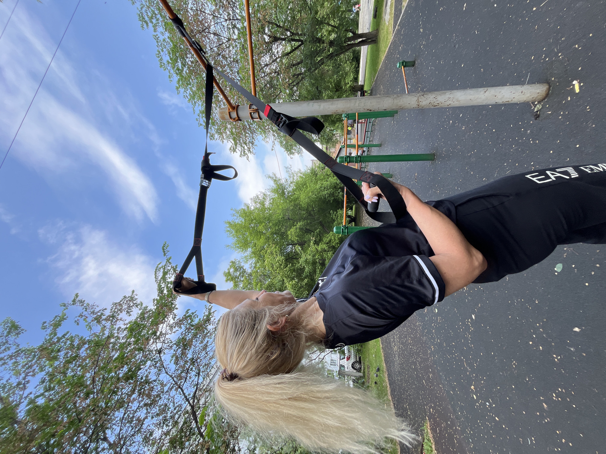 A woman with long blonde hair pulled back in a ponytail is performing an exercise with suspension trainers on an outdoor fitness equipment setup. She is wearing a black t-shirt with white trim and black pants with white lettering that reads "EA7 EMP". The fitness equipment includes bars and green posts, and the ground is a black rubberized surface. The scene is outdoors with trees and a cloudy blue sky in the background. There is a white car visible in the distance. This is an outdoor fitness area in the city of Krasnoyarsk, Russia.