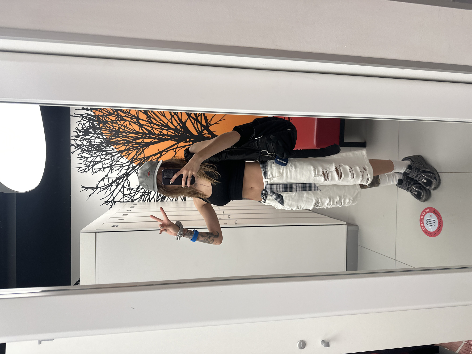 A person is taking a mirror selfie in what appears to be a locker room or changing area. The person is wearing a black tank top, white ripped denim shorts, a plaid belt, white socks, and dark sneakers. They are also wearing a gray baseball cap and have tattoos on their arm. They are holding up two fingers on their right hand and a phone in their left hand, with a black backpack slung over their shoulder. In the background, there are white lockers and a wall with a black tree silhouette against an orange background. A red bench is visible in the reflection. On the floor, there is a circular red and white sticker with text. The overall environment suggests a gym, spa, or similar facility. The sticker on the floor appears to have Cyrillic text and a symbol, potentially indicating a social distancing marker or a facility rule. There is no specific visual information in the image that definitively places this scene in Moscow, Russia.