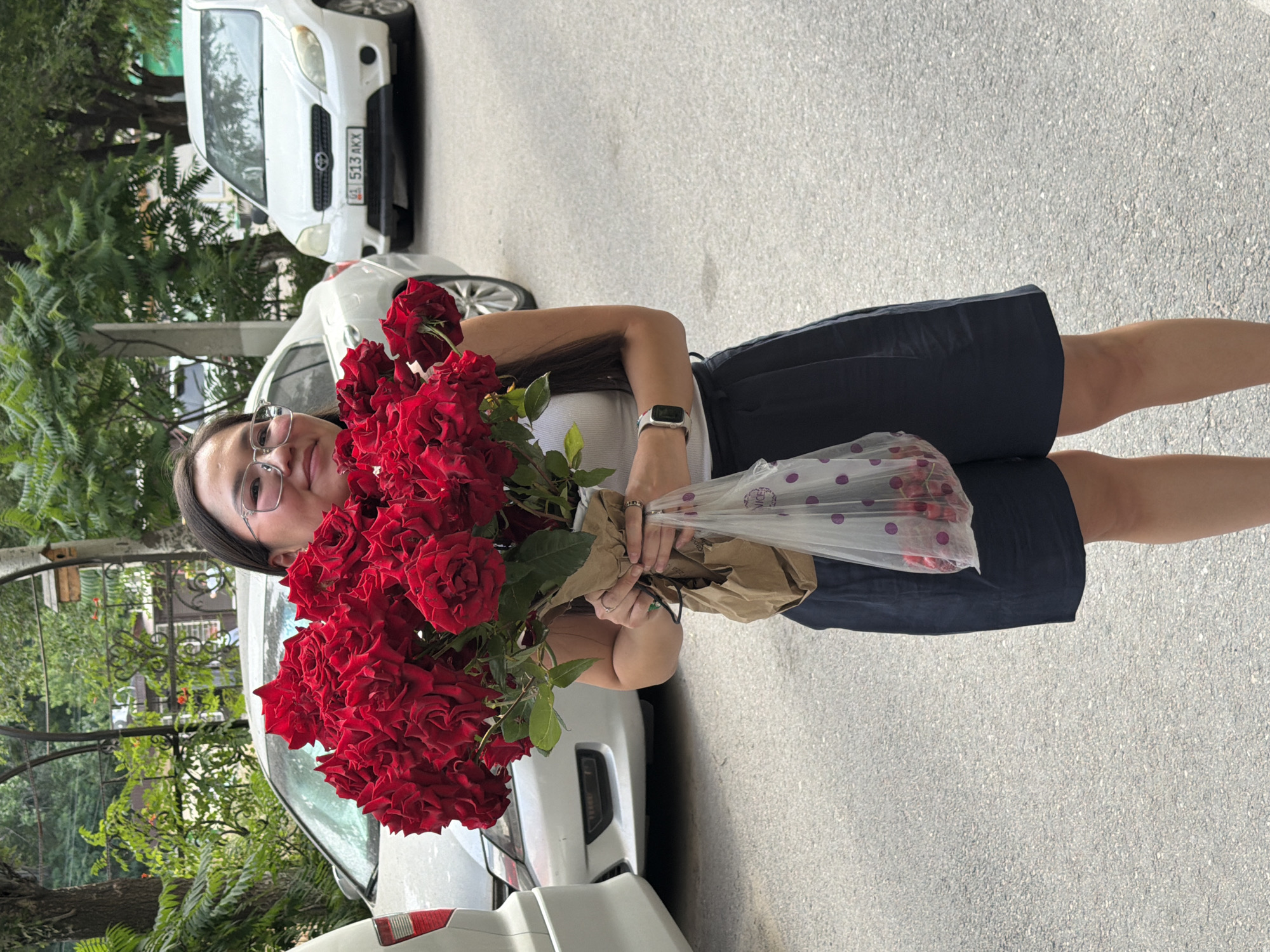 A young woman stands on a paved road, holding a large bouquet of red roses wrapped in brown paper. She is wearing a white tank top, dark blue shorts, and glasses. In her left hand, she holds a clear plastic bag filled with red cherries. Two white cars are parked behind her. The scene appears to be in an urban area, with trees and a decorative metal arch visible in the background.