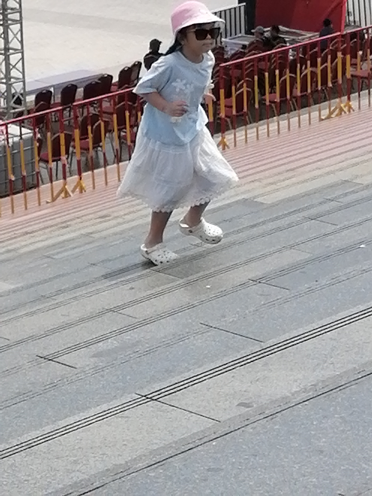 An outdoor, eye-level, medium shot shows a young girl wearing a pink bucket hat and sunglasses, running up a set of concrete stairs. She has on a light blue short-sleeved t-shirt with a white ruffled skirt and white crocs. The girl is in motion, with her arms bent at the elbow and her right leg extended upwards in a running stride. Behind her, a red metal railing with yellow vertical bars separates the stairs from a seating area with dark chairs. In the distance, more people are visible in the background, along with some structural elements, possibly part of a stadium or amphitheater. The concrete stairs have dark lines running across them, creating a textured surface. The scene appears to be a public space, possibly a park or a venue, in Da Lat, Vietnam, suggested by the style of the railing and the overall ambiance.