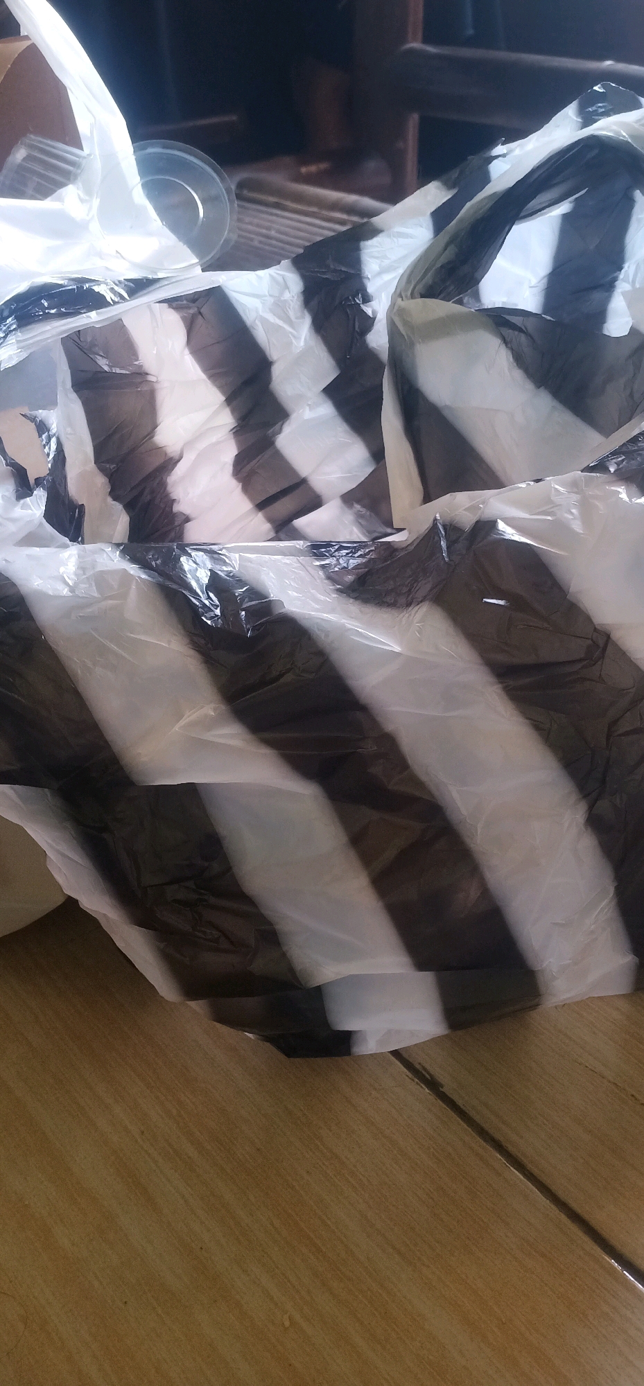 The image shows a pile of crumpled plastic bags with a black and white striped pattern, lying on a wooden floor. Some of the bags are translucent white, while others are opaque black, creating a diagonal striped effect. A clear plastic cup is visible on top of one of the bags, and a ribbed object, possibly part of a fan or another appliance, is partially visible in the background. The overall scene appears to be indoors, possibly in a home or storage area, and does not provide specific location details within Bajong, Indonesia. There are no people or any discernible actions taking place. The lighting is dim, with shadows cast by the objects.