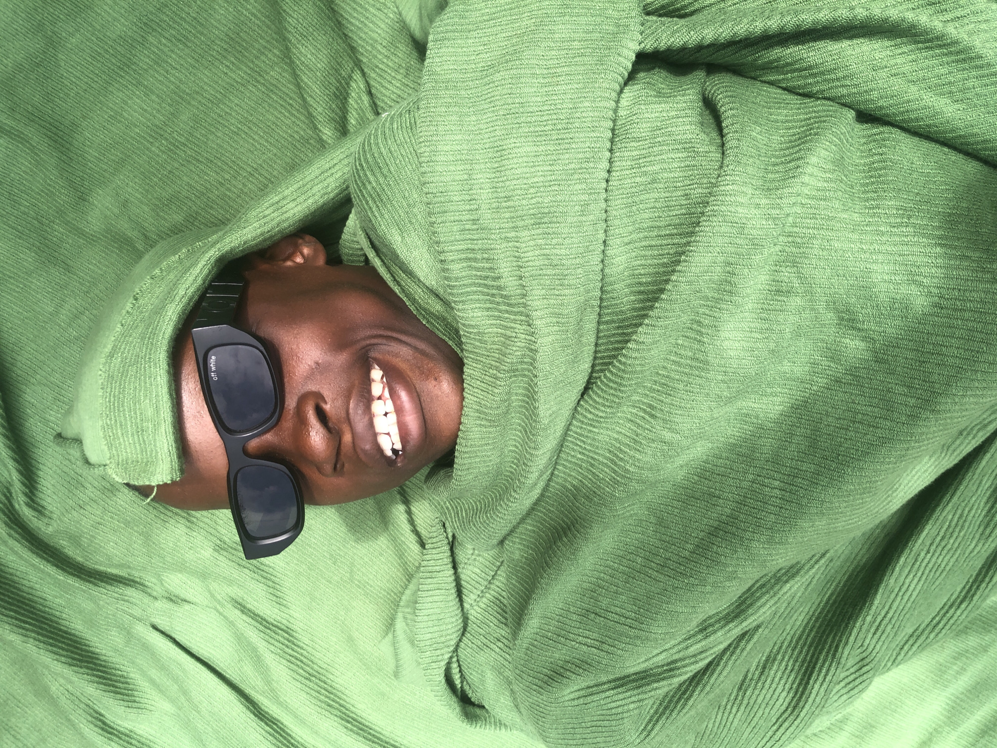 A young Black person with dark skin, wearing black "Off-White" sunglasses and a green hooded garment, lies on a textured green fabric surface. The person's eyes are visible through the sunglasses, and their mouth is open, showing white teeth. The green fabric appears to be a soft, textured material, possibly corduroy or a similar ribbed fabric, and it is draped around the person, partially obscuring their body. The overall impression is of a person relaxing or posing within the fabric. The background is entirely composed of this green fabric.