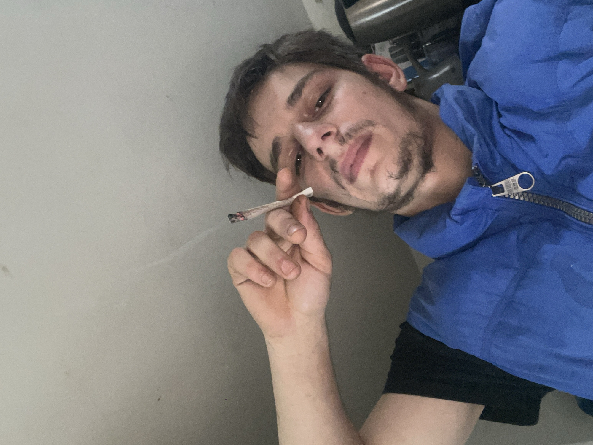 A man is holding a lit cigarette. He is wearing a blue jacket and black shorts. The location appears to be indoors.