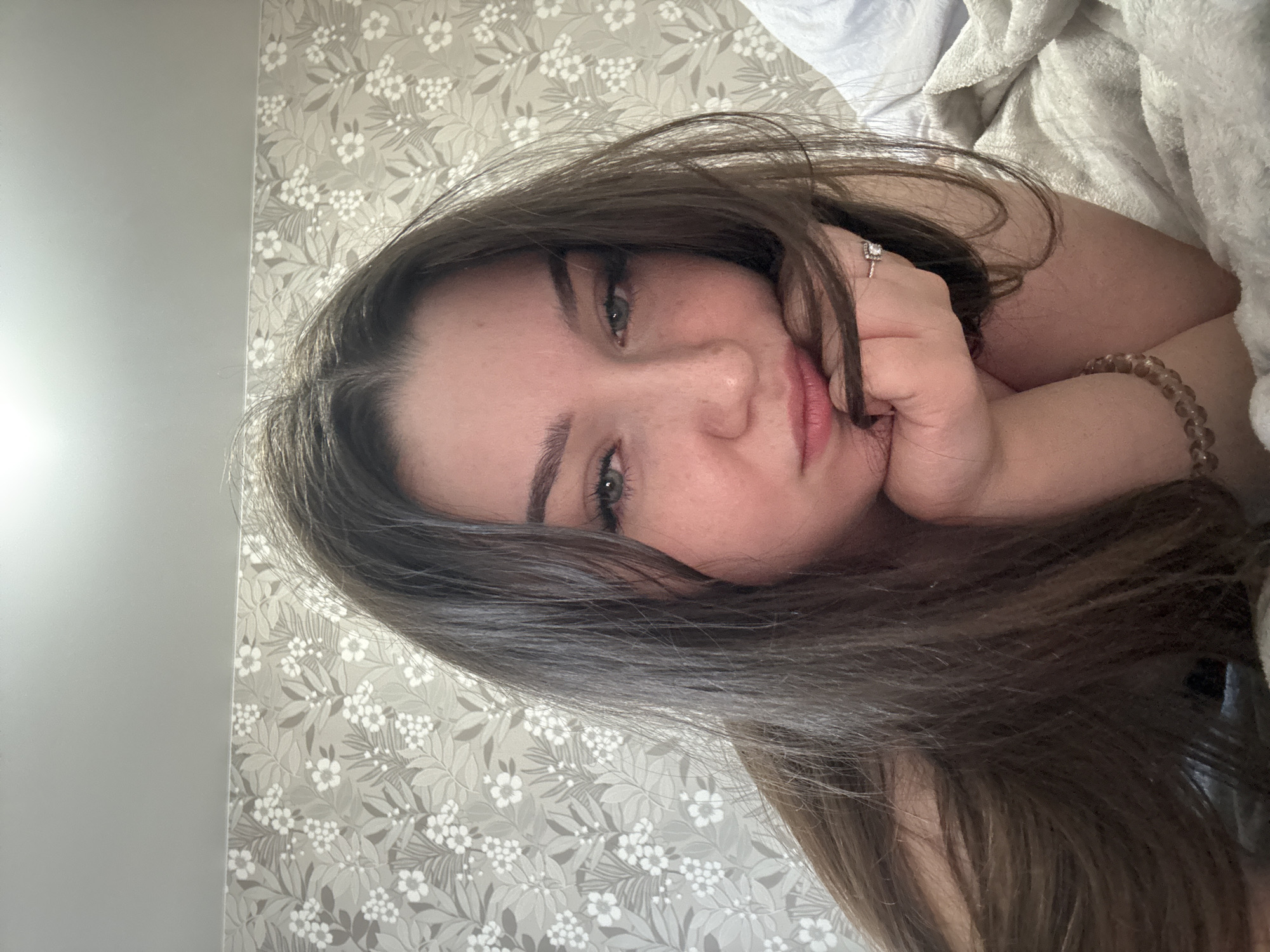 A young woman with long brown hair is lying on a bed, propped up on her arms. She is looking at the camera with a neutral expression. The background is a wall with floral wallpaper. The lighting suggests it is daytime. There are no indications of the location being in Zarechnaya (Gamovskoe S/P), Russia.