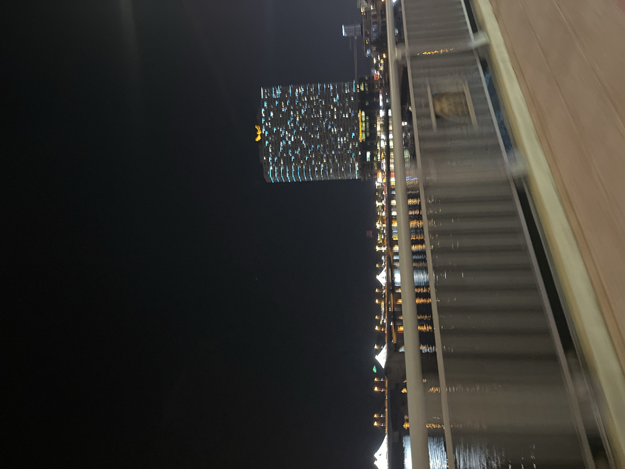 The image shows a nighttime view in Bac Nha Trang Ward, Vietnam. A tall building with numerous illuminated windows stands out against the dark sky. Reflections of lights shimmer on the water below. The photograph seems to be taken from an elevated position, possibly from another building or bridge, with a portion of the structure visible in the foreground on the right. The foreground structure has horizontal lines and a beige color to it.