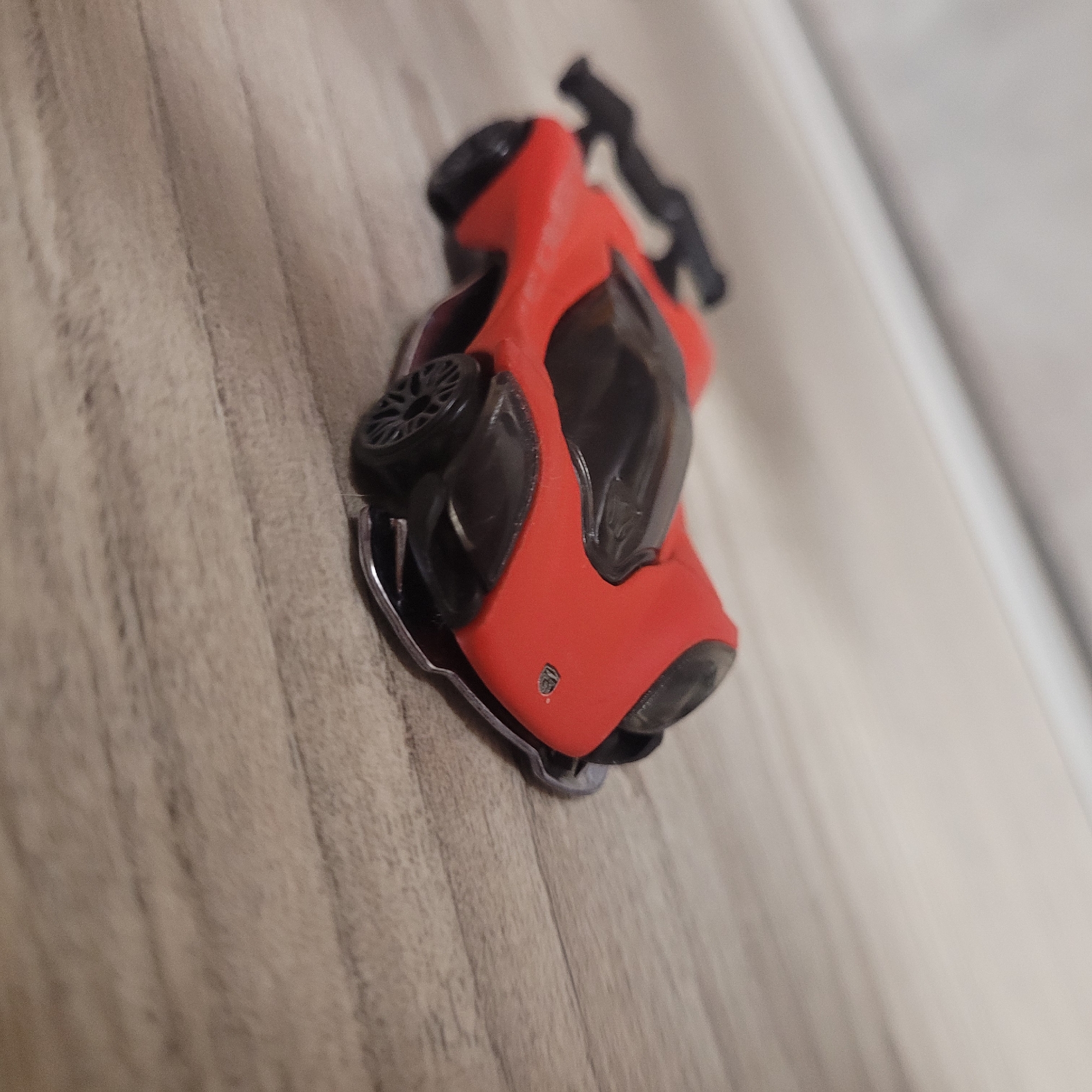 A red toy car is resting on a wooden surface. The car has black accents and a black spoiler. The surface appears to be a table or floor with wood grain. The background is blurred and out of focus, showing a light-colored wall or surface. The lighting suggests indoor conditions.