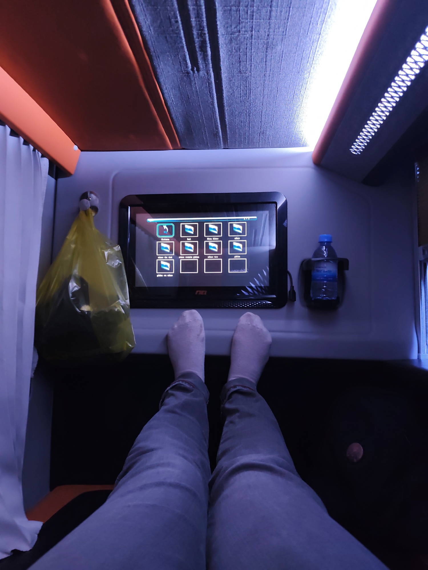 The image captures a first-person perspective from inside a bus or train compartment, likely a sleeper or first-class seat. The camera is positioned low, looking up at a screen and further up at the ceiling. The foreground shows a person's legs and feet, clad in light-colored socks and gray pants, extending into the frame. The compartment is dimly lit, with a blue-toned ambient light reflecting off surfaces.
A prominent feature is a tablet-like screen mounted on the wall in front of the seat, displaying a menu with several icons, possibly for entertainment or services. To the left of the screen, a yellow plastic bag, possibly containing items, is hanging from a hook. To the right of the screen, a bottle of water in a black holder is attached to the wall.
The ceiling of the compartment is textured and has integrated lighting, with orange accents along the edges. White curtains are visible on the left side, likely covering a window or partition. The overall impression is one of a private, modern transportation environment designed for comfort and entertainment during travel. The exact location is unknown.
