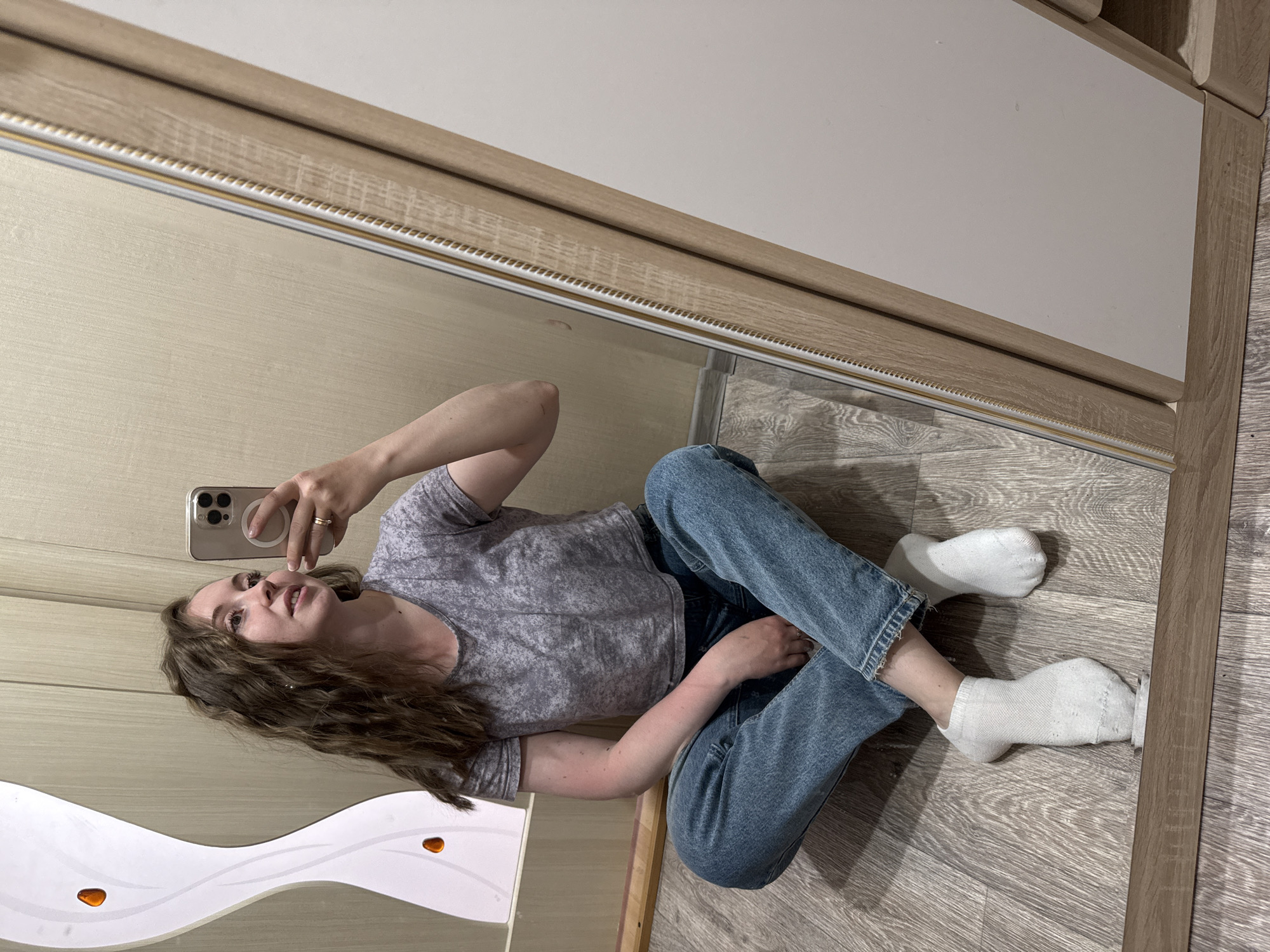 A woman wearing a gray t-shirt, blue jeans, and white socks is lying on a wooden floor, taking a selfie in a mirror. The mirror is framed by wooden trim. A white wavy-shaped object with amber-colored decorations is in the reflection, located to the woman's left. The location is inside a room with wooden flooring and furniture. The city and location are unknown.