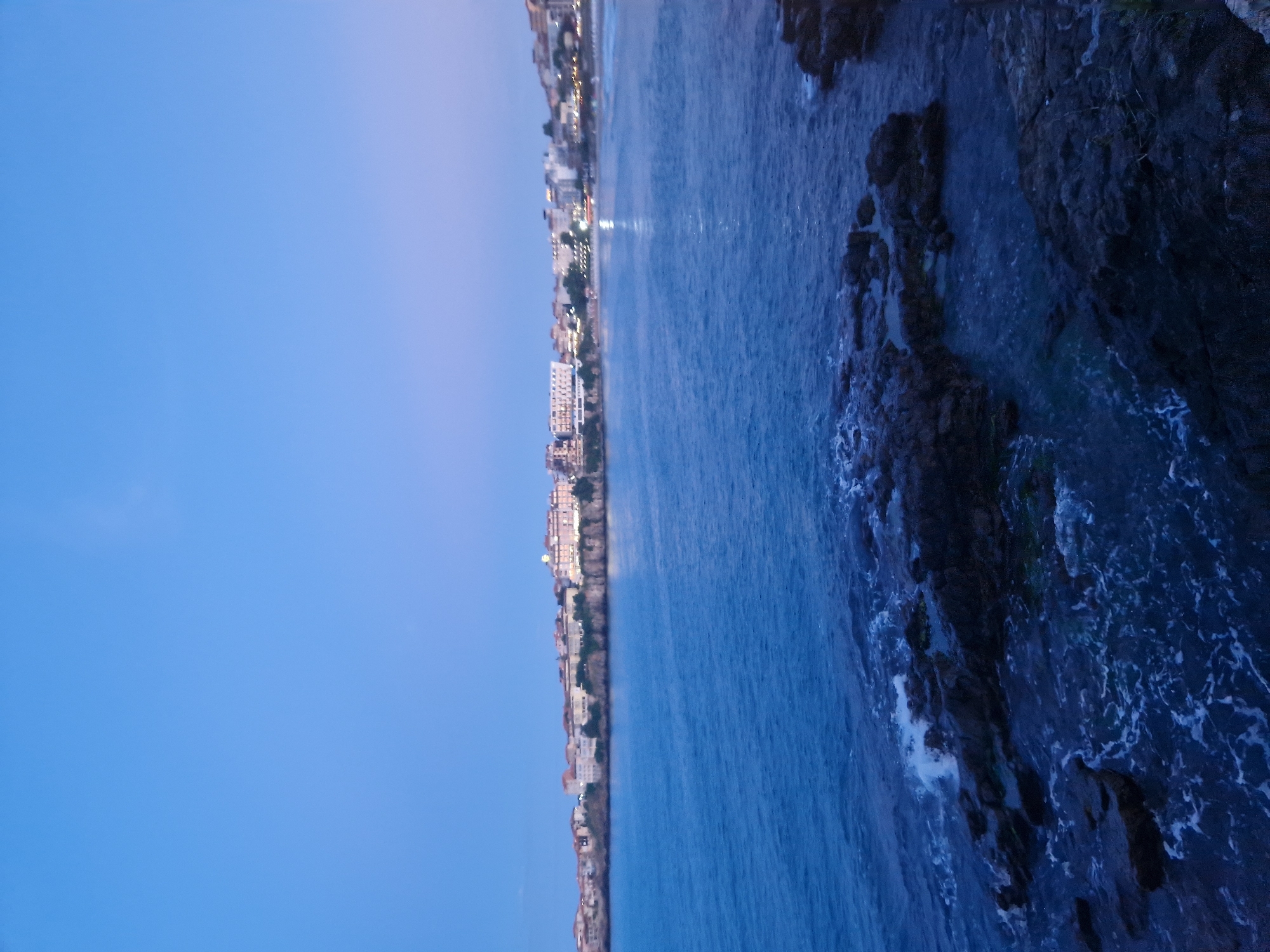 An exterior, low-angle, wide shot captures the coastline of Sozopol, Bulgaria, at twilight. The sky is a deep, gradient blue, transitioning to a lighter, almost purple hue near the horizon. The sea, a dark blue, stretches across the lower half of the frame, with gentle waves lapping against rocky outcrops in the foreground. White foam churns around the dark, wet rocks, creating a stark contrast.
In the distance, a long line of buildings, likely hotels and houses, forms the city skyline. The architecture appears to be a mix of styles, with some structures featuring tiled roofs and others more modern facades. Lights are beginning to twinkle from within the buildings, indicating that the evening is settling in. A beach is visible in the far distance, with faint lines suggesting palm trees or umbrellas. The overall atmosphere is tranquil and serene, evoking the peaceful end of a day in a coastal town. No people are visible in the image.