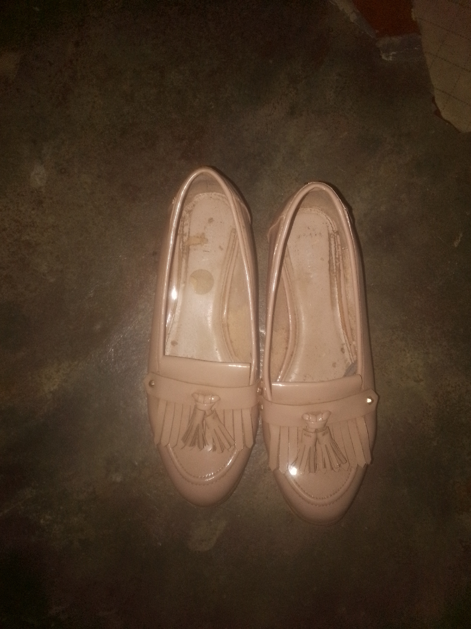 Two light-pink loafers are positioned side-by-side on a dull, dark-brown surface in Sabon Tasha, Nigeria. The loafers, identical in design, feature a rounded toe and a fringe detail. They show signs of wear.