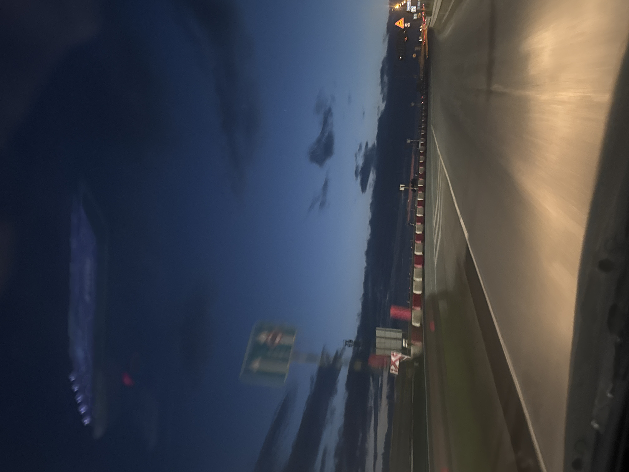 The image shows a view from a vehicle moving along a road at dusk or dawn. The sky is a deep blue with scattered clouds, suggesting either early morning or late evening. The road itself is wet, reflecting the artificial lights from overhead lamps and road markers, indicating recent rain or high humidity.
Along the side of the road, there are red and white striped barriers, typical of road construction or safety zones. Further down the road, signs are visible, though their text is blurred due to motion and distance. One sign appears to be a triangular warning sign with a yellow background, commonly used for road hazards. Another sign, larger and green, is also present, likely indicating directions or information about the route.
In the distance, the lights of what appears to be a settlement or more distant traffic are visible. The overall atmosphere is one of travel and transition, with the fading light and wet road contributing to a somewhat subdued mood. The specific location is identified as Kondratovo, Russia, which provides a geographical context to the scene. There are no discernible people in the image. The primary focus is the road and its surrounding environment during low light conditions.