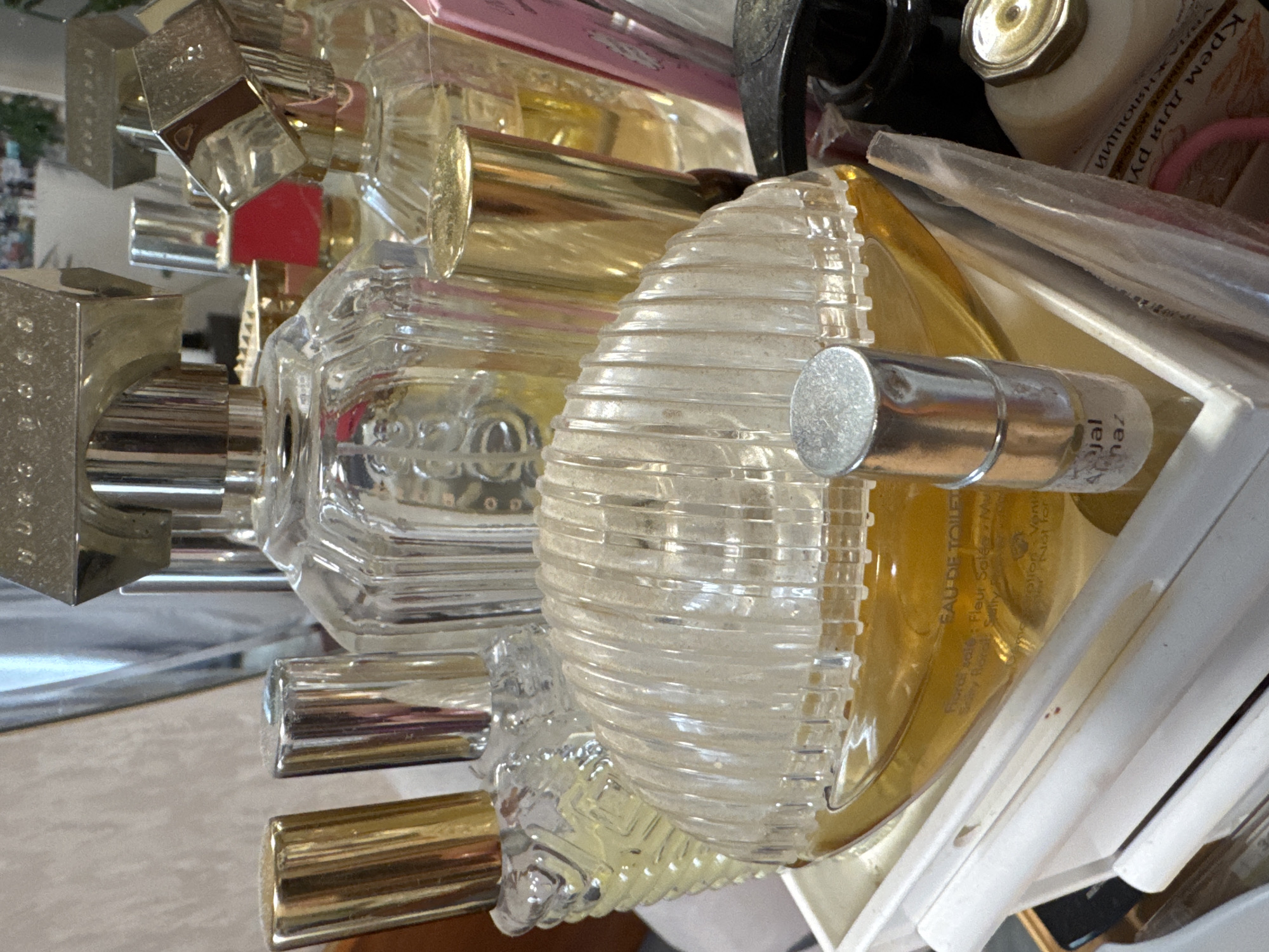 This media file captures a close-up, overhead view of a cluttered collection of personal care products, likely on a shelf or vanity. The scene is brightly lit, indicating an indoor setting.
The primary subjects are numerous perfume bottles and cosmetic items. Several glass perfume bottles are visible, with varying shapes; one prominent bottle features a distinctive ribbed, clear glass design. Many bottles have metallic caps or spray components in silver and gold tones. One square, silver-capped bottle clearly bears the brand name "HUGO." Another silver cylindrical bottle, possibly a travel-sized perfume or roll-on, is labeled "Ajmal Almaz." A white bottle with a gold-colored cap is also present, identified by its Cyrillic text as "Крем для рук" (Hand Cream) and "УВЛАЖНЯЮЩИЙ" (Moisturizing).
Visible text also includes "EAU DE TOILE" (Eau de Toilette), "Fioral sale," "Fleur Salée M," and "Plion Vente," possibly related to a specific fragrance name or type. The partial phrase "Not for" is also seen. The presence of Cyrillic text on the hand cream bottle is consistent with the location context of Izhevsk, Russia. The items appear somewhat disorganized, suggesting a lived-in space rather than a retail display. Some surfaces show light dust, particularly on the ribbed glass bottle. No people or activities are depicted, focusing solely on the objects.