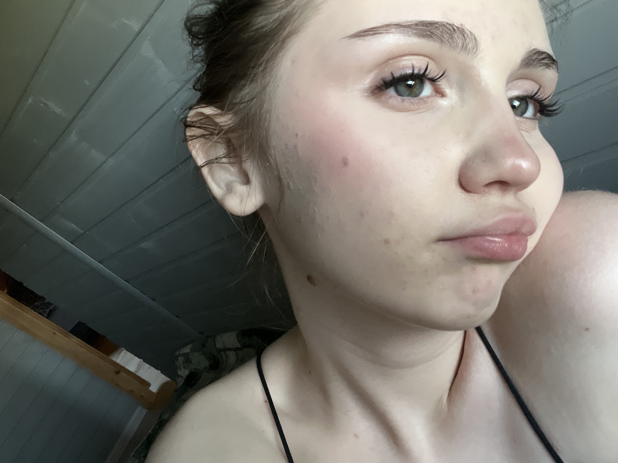 This is a close-up, high-angle medium shot of a young woman's face, with her head turned away from the camera and her gaze directed off to the right. The woman has light skin with some visible blemishes and redness on her cheeks. Her eyebrows are dark and well-defined, and her eyelashes are long and dark. Her eyes are a light green or grey color. Her lips are full and slightly pursed, with a glossy sheen. She appears to be wearing subtle makeup, with a hint of blush on her cheeks and possibly mascara and lip gloss. Her hair is dark and pulled back in a messy bun.
The background is out of focus but appears to be indoors, with a wall composed of horizontal wooden planks painted a dark grey. To the left, a lighter wooden structure, possibly part of furniture or a shelf, is visible. The lighting suggests it is daytime, possibly late afternoon or early evening due to the warm tones on her skin. The overall mood is casual and intimate. There is no visible text in the image.