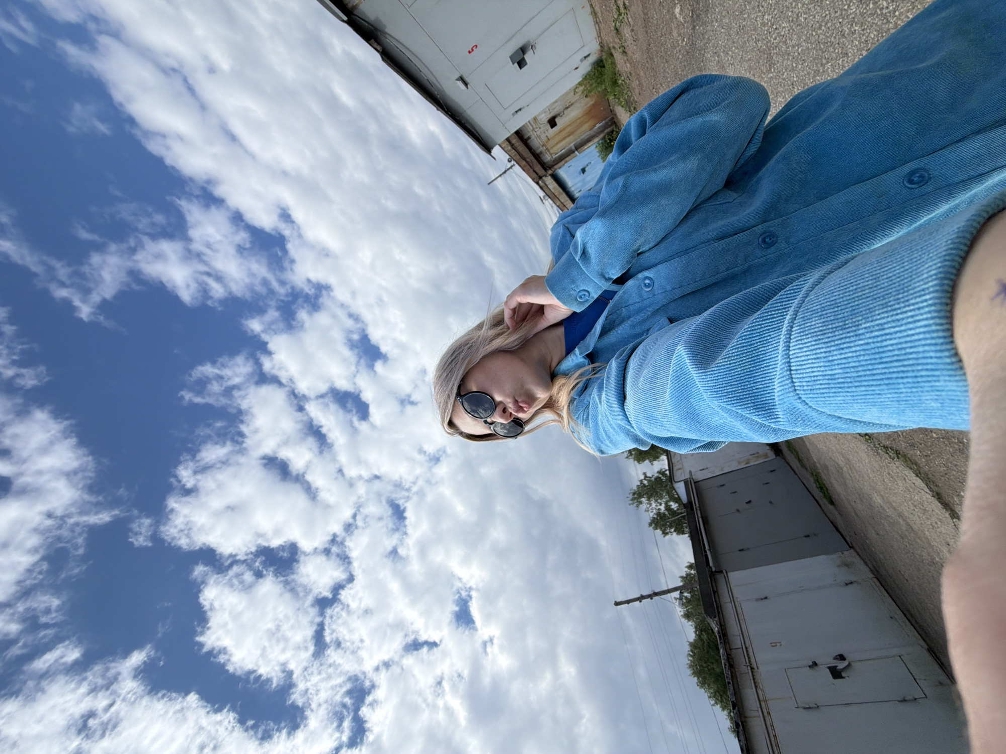 This tilted, low-angle shot captures a young woman posing outdoors against a backdrop of garages and a partly cloudy sky. The woman, with blonde hair, is wearing dark sunglasses and a textured, vibrant blue corduroy shirt or jacket. She looks towards the right side of the frame with a slightly puckered lip expression, possibly adjusting her hair or sunglasses with her left hand.
The upper and central parts of the image are dominated by the sky, which features both clear blue patches and scattered white, wispy clouds. In the background, a line of grey, weathered garage doors is visible, suggesting an urban or suburban setting. The number '5' is clearly marked in red on one of these garage doors. A utility pole and some green foliage, possibly trees or bushes, can also be seen. The ground in the foreground and along the garages appears to be a paved surface like asphalt or concrete. The image portrays a casual, daytime moment in what appears to be an alleyway or back street in Chaikovsky, Russia.