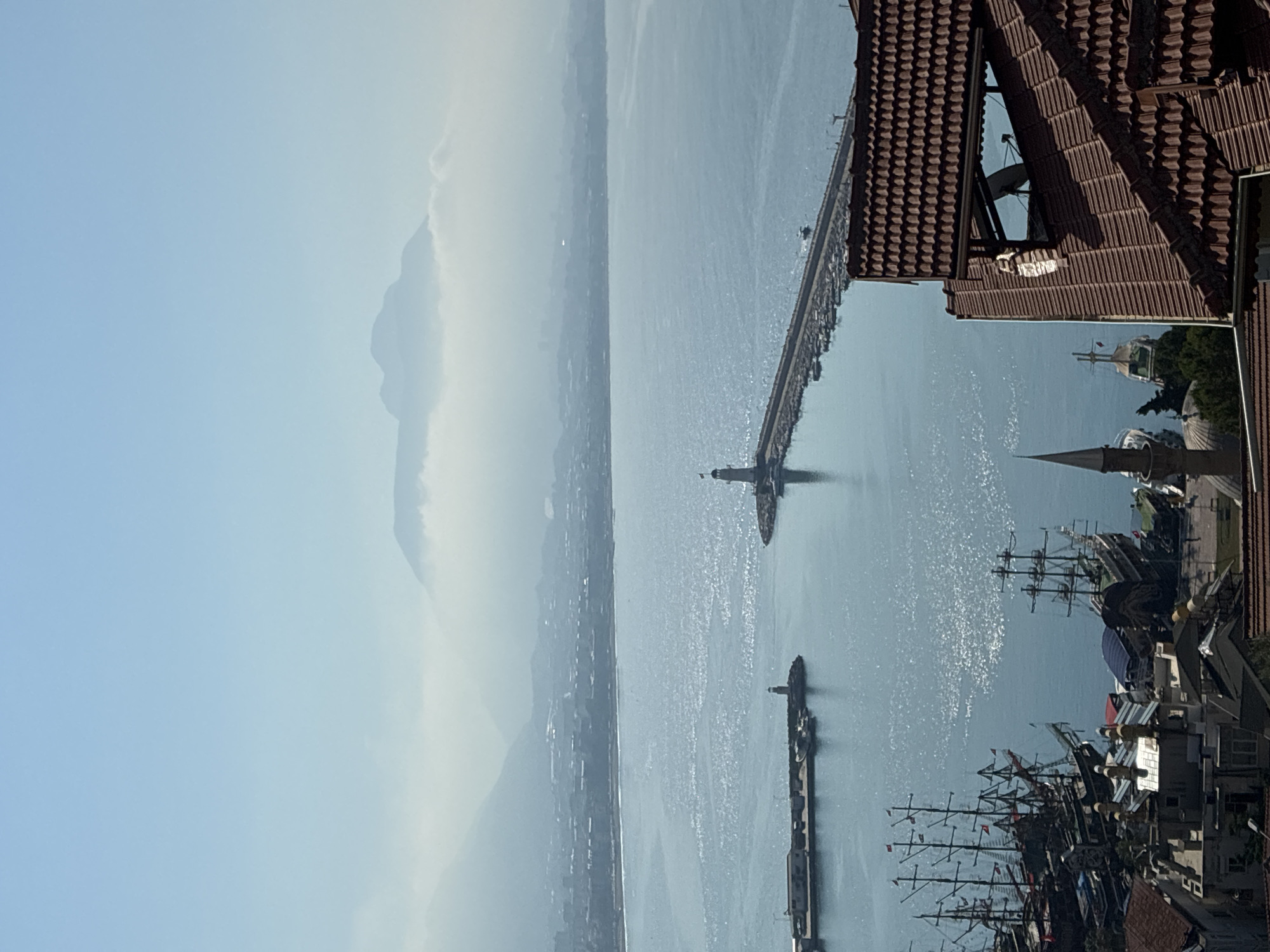 The image is a high-angle, wide shot of Alanya, Turkey, looking out over the harbor towards the mountains in the distance. The sky is clear and blue, with a light haze obscuring the far mountains. In the foreground, a cluster of red-tiled roofs belonging to buildings in the city are visible.
The harbor itself is a prominent feature, with a long breakwater extending into the sea, topped with a lighthouse at its end. Numerous boats, including several large, traditional-style wooden sailing ships, are moored along the breakwater and closer to the shore. The water in the harbor is a deep blue, reflecting the sunlight with a shimmering effect.
In the middle ground, a strip of coastline with buildings stretches across the horizon. Beyond the city, the hazy mountains rise, with wisps of clouds clinging to their peaks. The overall impression is one of a sunny day in a coastal city, with a bustling harbor. There are no visible people or explicit actions taking place. The focus is on the landscape and the maritime elements of Alanya.