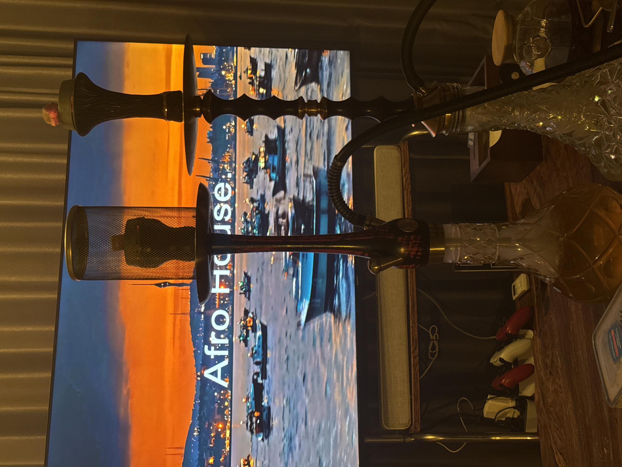 The image depicts an indoor scene, located in North Kuta, Indonesia, characterized by dim, warm artificial lighting and the glow of a television screen. In the foreground, two hookahs stand on a dark wooden surface.
The hookah on the left has a dark, patterned stem and a clear, faceted glass base containing a light brown liquid. A cylindrical, black mesh heat management device, with a dark object visible inside, rests on top. A black hose extends from the base.
The hookah on the right features an ornate, dark-toned stem with visible texture and gold-colored accents, positioned above a clear, faceted glass base containing a clear liquid. A dark bowl, topped with a reddish, granular substance, is visible. A black hose is coiled on the right side of the hookah, and a small, round wooden cap is situated next to its base.
A large television screen dominates the background, displaying a panoramic scene of a vibrant sunset or sunrise over a body of water. The sky transitions from a deep orange and red at the horizon to a dark blue hue at the top. Numerous small to medium-sized boats are scattered across the water. A city skyline with illuminated buildings and several tall, slender structures, including one resembling the Istanbul TV Tower, are silhouetted against the bright sky. The text "Afro House" is displayed prominently in large white letters across the lower portion of the screen.
Below the television, a light-colored soundbar is centered on a wooden shelf. To the left of the hookahs, on the same shelf, a white power strip is visible, along with three charging devices resembling electric clippers or shavers (two red, one off-white), and a small, light-colored remote control. Various cables and wires are present behind these items. To the right, a small wooden box and a clear glass jar with a wooden lid are partially visible. The room's backdrop consists of dark brown, possibly pleated, curtains. No individuals are present in the image.