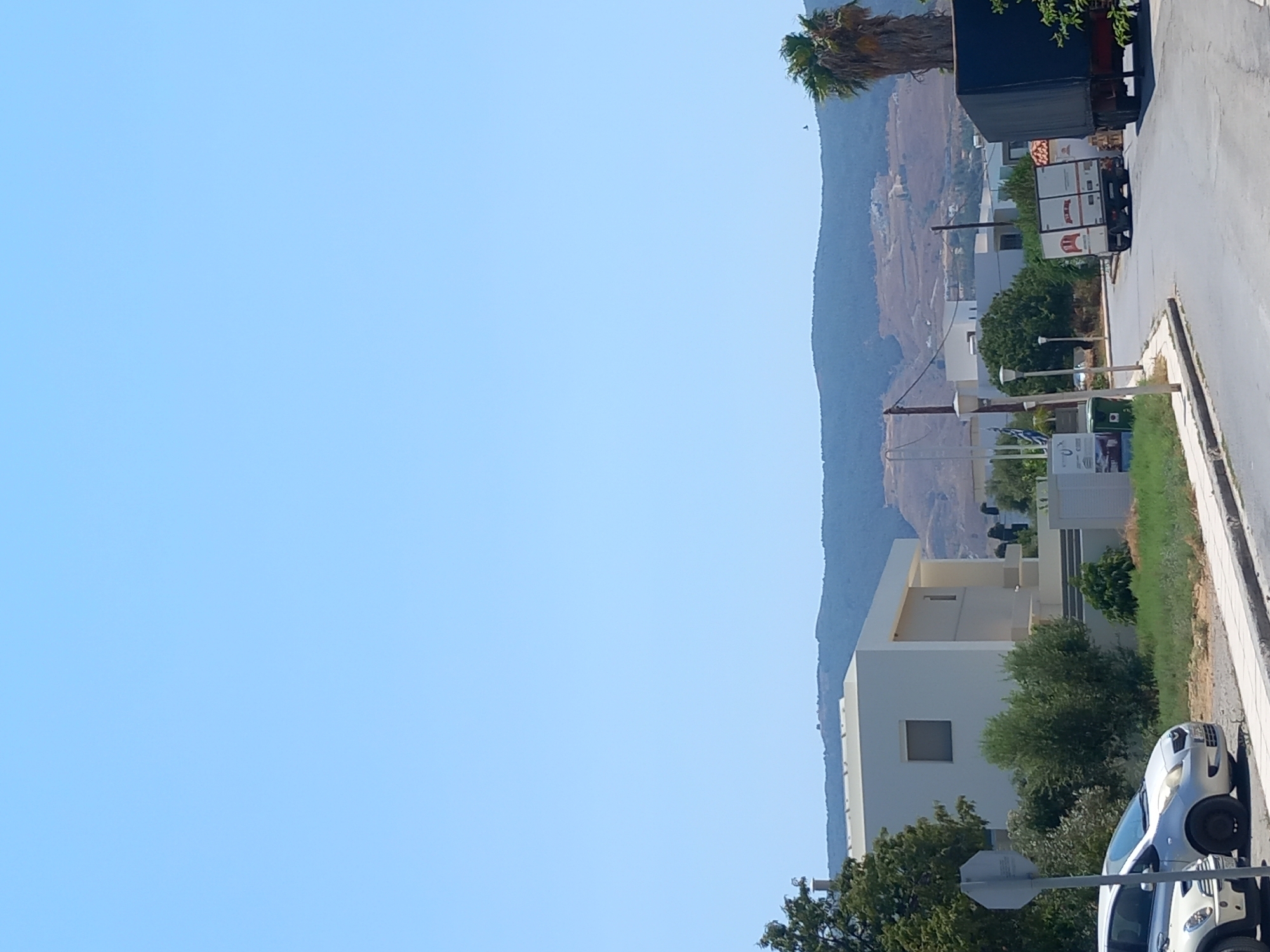 In Kos, Greece, a bright sunny day is captured with a clear blue sky. The scene features a modern white building with clean lines and a flat roof. In the foreground, two cars are parked on the left side of a paved road. One is a silver hatchback, and the other, partially visible, is a white compact car. A gray traffic sign is positioned on the left, near the parked cars.
Further down the road, a small white truck with a red logo is visible, parked at an angle. To the right of the truck, a dark blue tarpaulin covers a trailer, suggesting a larger vehicle parked nearby, possibly a truck. A tall palm tree stands on the right side of the image.
In the background, rolling hills are covered with sparse vegetation and some scattered buildings, indicating a semi-urban or rural outskirts setting. A winding road is visible on the hillside. The overall atmosphere is one of a typical day in a Mediterranean locale.