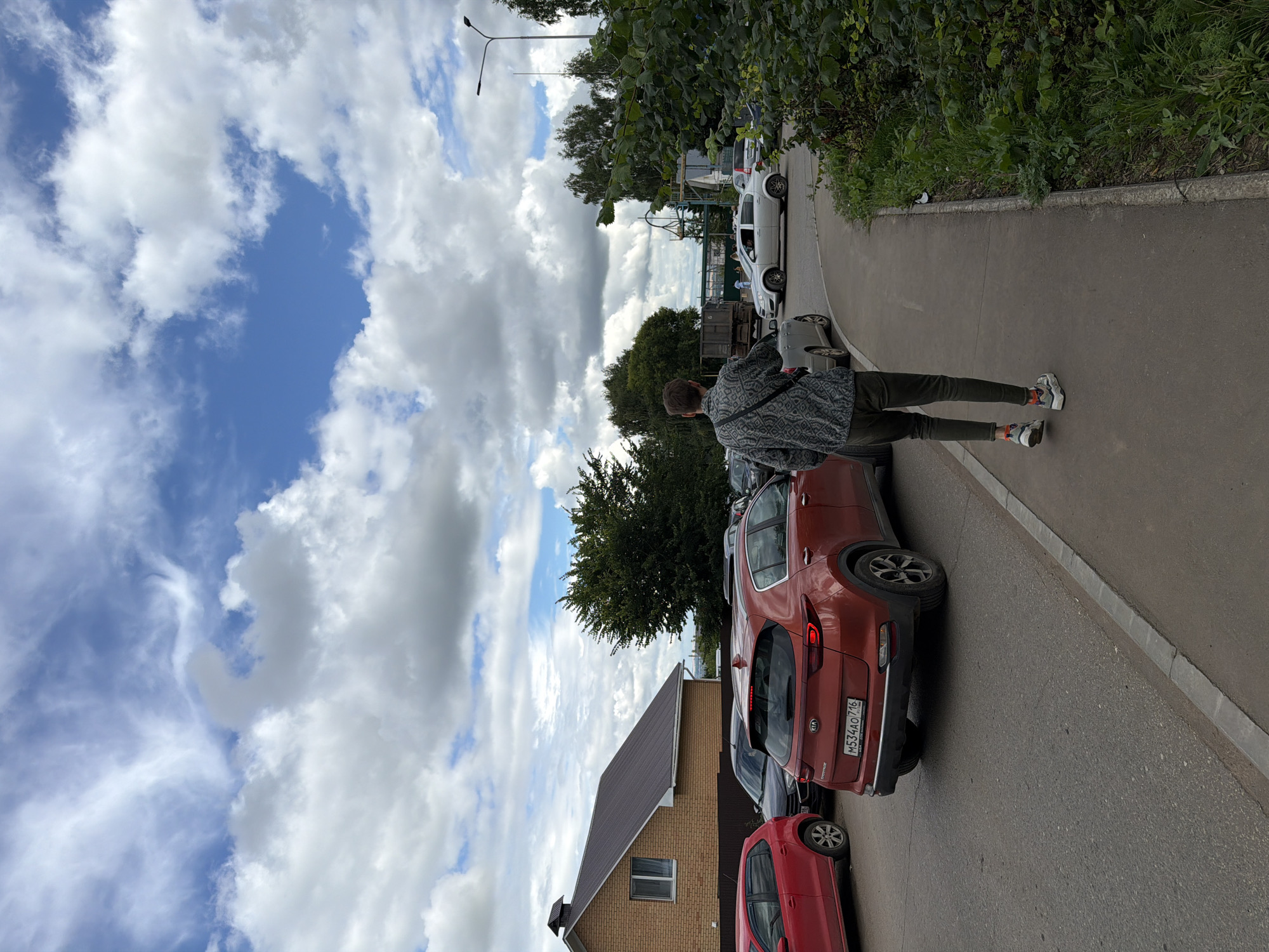 In Yelabuga, Russia, on a cloudy day, a young man in a patterned shirt and dark pants walks away from the camera on a paved sidewalk. He has a dark bag slung across his chest. The sidewalk is bordered by grass and vegetation on the right. A road runs parallel to the sidewalk on the left, with several cars stopped in traffic. Among them is a red Kia Sportage with a license plate reading M534AO 716. Further down the road, a silver car is making a turn. In the background, there is a brick house with a dark roof, trees, and a lamppost. The sky is filled with dramatic clouds.