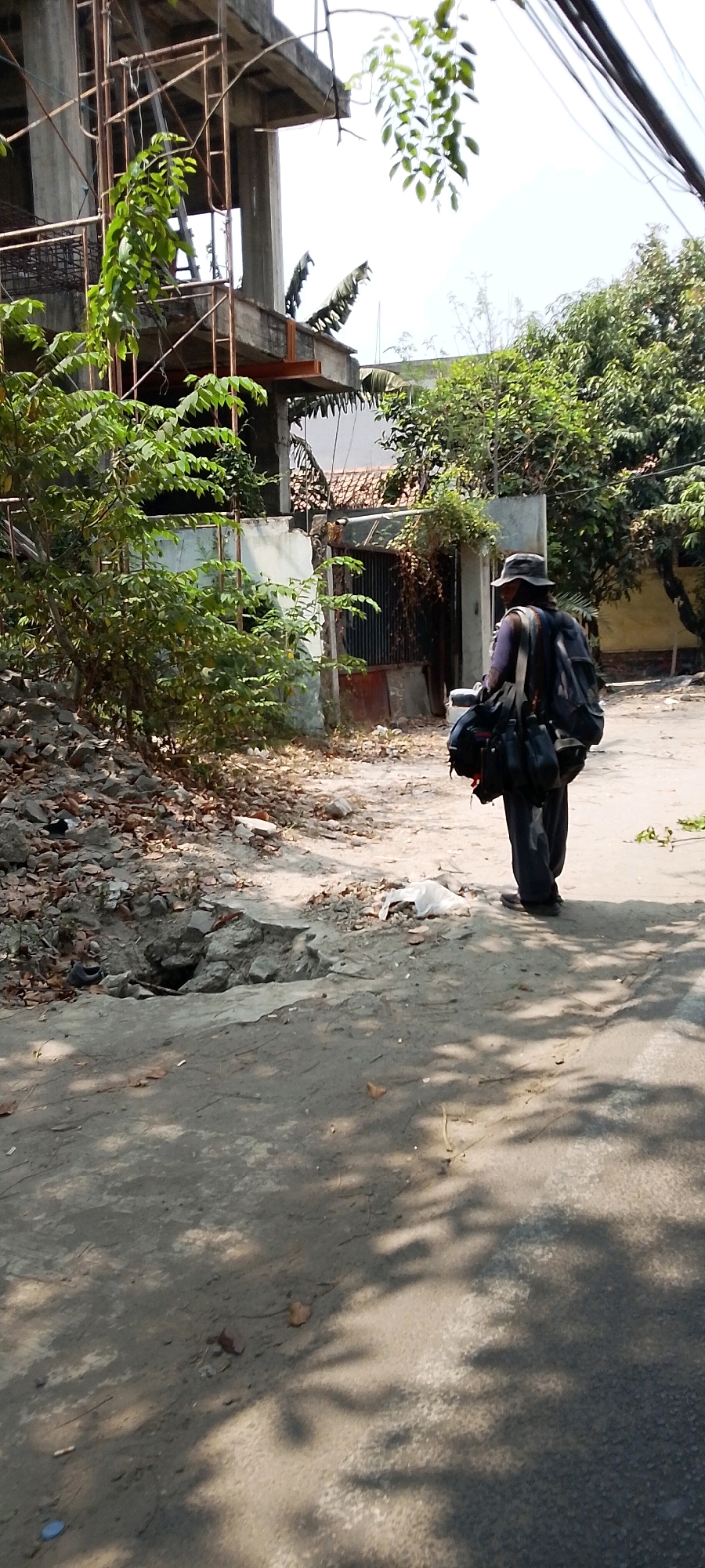 In Bekasi, Indonesia, a person wearing a hat and carrying a large backpack walks on a dirt road. The person is facing away from the camera and is seen from the back. The road has some debris and a hole in the ground. Behind the person, there is an unfinished building with scaffolding and some trees. The sky is bright and clear, suggesting a sunny day.