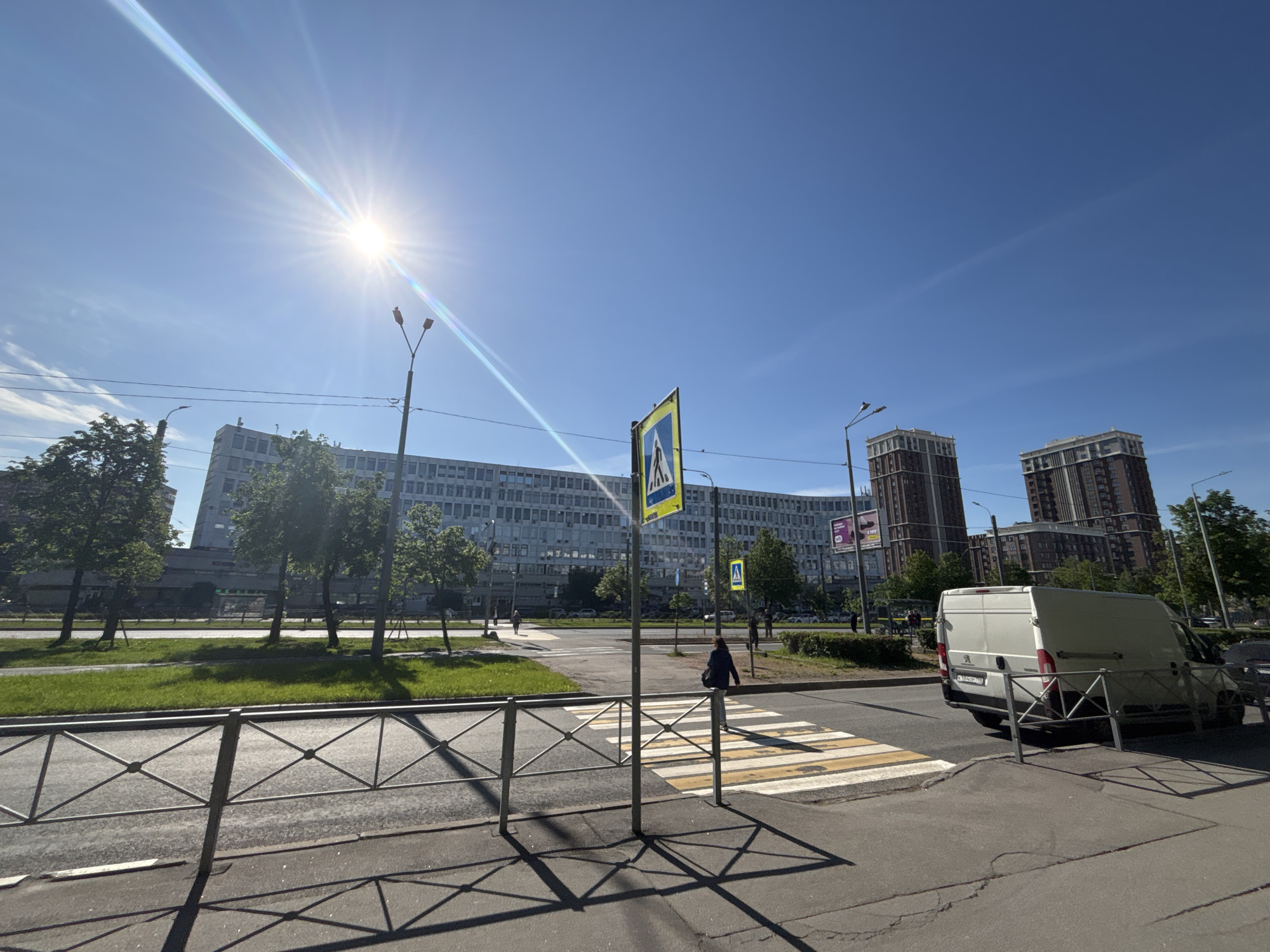 The image presents a bright, sunny daytime street scene in an urban or suburban setting, likely in Russia, indicated by a visible license plate. The sky is clear and vivid blue, with a strong sunburst and lens flare emanating from the upper left.
In the immediate foreground, a metal pedestrian barrier fence casts distinct, geometric shadows across the pavement. A prominent zebra crossing (pedestrian crosswalk) is marked with yellow and white stripes, and an associated pedestrian crossing sign (blue square with a white triangle and walking figure) stands beside it. A person is visible near this crosswalk.
The midground features a multi-lane road with a grassy median strip lined with trees. On the left, a long, multi-story building with numerous windows stretches across the frame. On the right side of the road, a white panel van, resembling a Peugeot, is parked or stopped, displaying a Russian license plate reading "C843OP 198".
In the background, two tall, modern residential buildings with unique architectural details at their tops rise against the horizon on the right. More trees and distant structures are also visible. Streetlights are present throughout the scene. A partially visible billboard-like sign on a distant building to the left shows the text "3 199". The overall impression is one of a bustling urban environment under clear weather conditions.