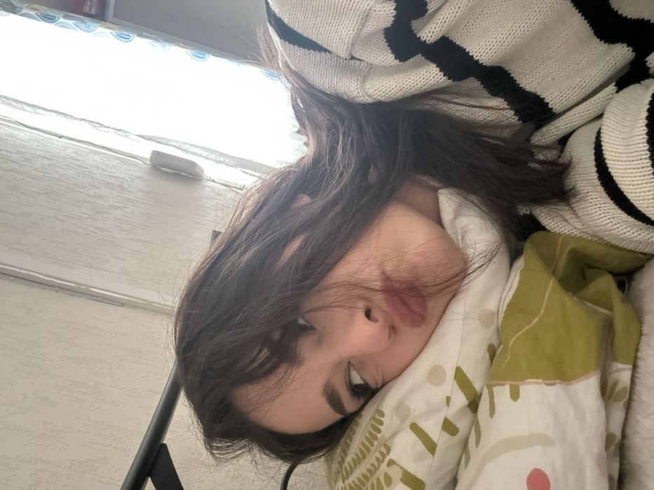 The image depicts a young adult female lying on a pillow, viewed from a close-up, slightly elevated perspective. She has long, dark hair, with some strands falling across her forehead and face. Her head rests on a white pillow adorned with a green and white abstract pattern, possibly depicting leaves or organic shapes. She is wearing a light-colored (likely white or off-white) knitted sweater with horizontal black stripes. Her facial expression appears contemplative or slightly pouting, with her lips pursed and her gaze directed towards the upper left side of the frame, away from the camera.
In the background, a light-colored wall with a subtle textured pattern is visible. To the right, a bright window is partially visible, allowing strong light to enter the room. A light-colored curtain or blind with a faint blue pattern (possibly teardrop or paisley shapes) partially obscures the window. A dark, possibly metal, bed frame or headboard element is visible in the upper left corner. The scene appears to be an indoor, domestic setting. The specific location within Strasbourg, France, cannot be determined from the visual information present in the image.