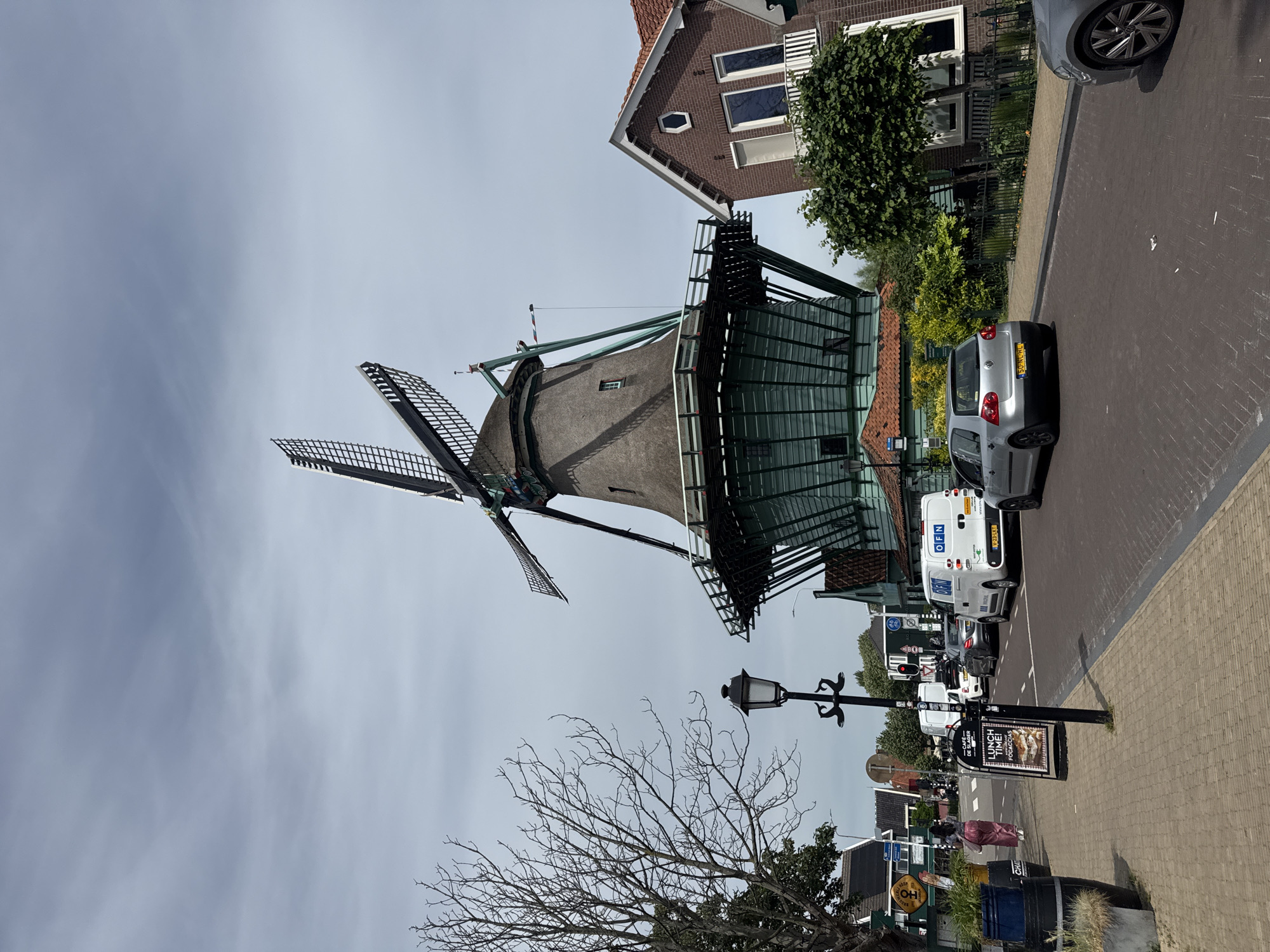 The image depicts a windmill in Zaandijk, Netherlands. The structure stands prominently, with its sails extending against the sky. Adjacent to the windmill is a building with a brick facade and visible windows. A street runs alongside the windmill, featuring parked cars and a white van with a visible license plate. A lamp post is also visible. A sign advertising lunch is seen. A bare tree stands in the foreground. Paved street is seen. Pedestrians walking in the background.