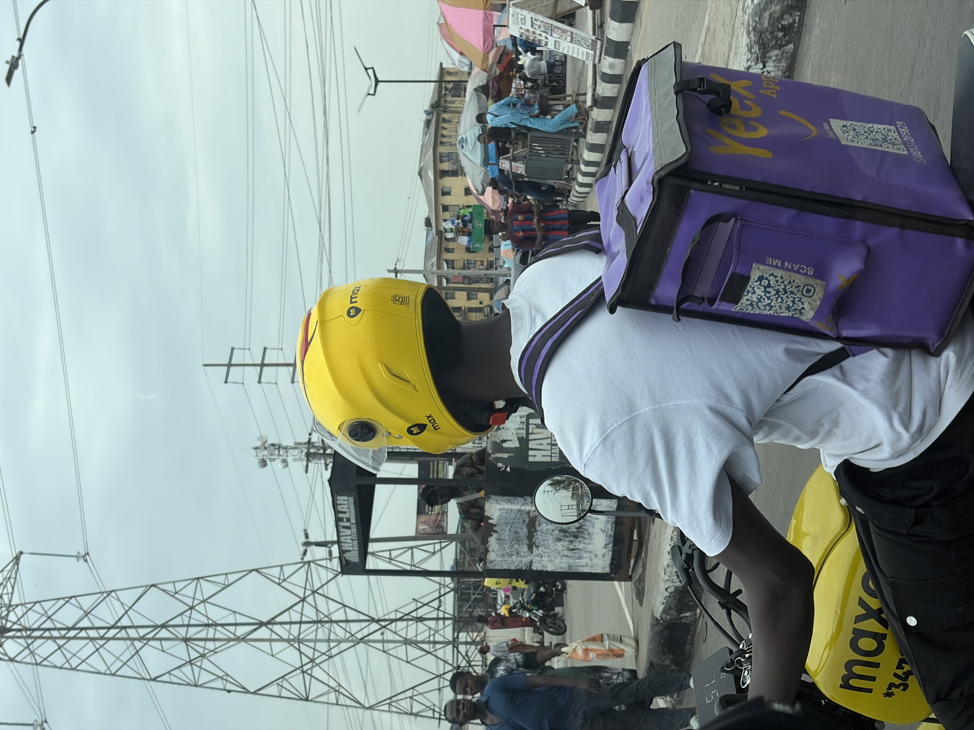The image captures a motorcyclist wearing a yellow helmet and a white t-shirt. Behind him is a purple delivery bag with "Yeek" and "App" printed on it, along with a QR code labeled "SCAN ME". The motorcycle is yellow and bears the word "max". The setting appears to be Igboefon, Nigeria, as evidenced by the roadside structures, utility poles, and general urban environment. People are visible in the background, along with various roadside stalls or shops.