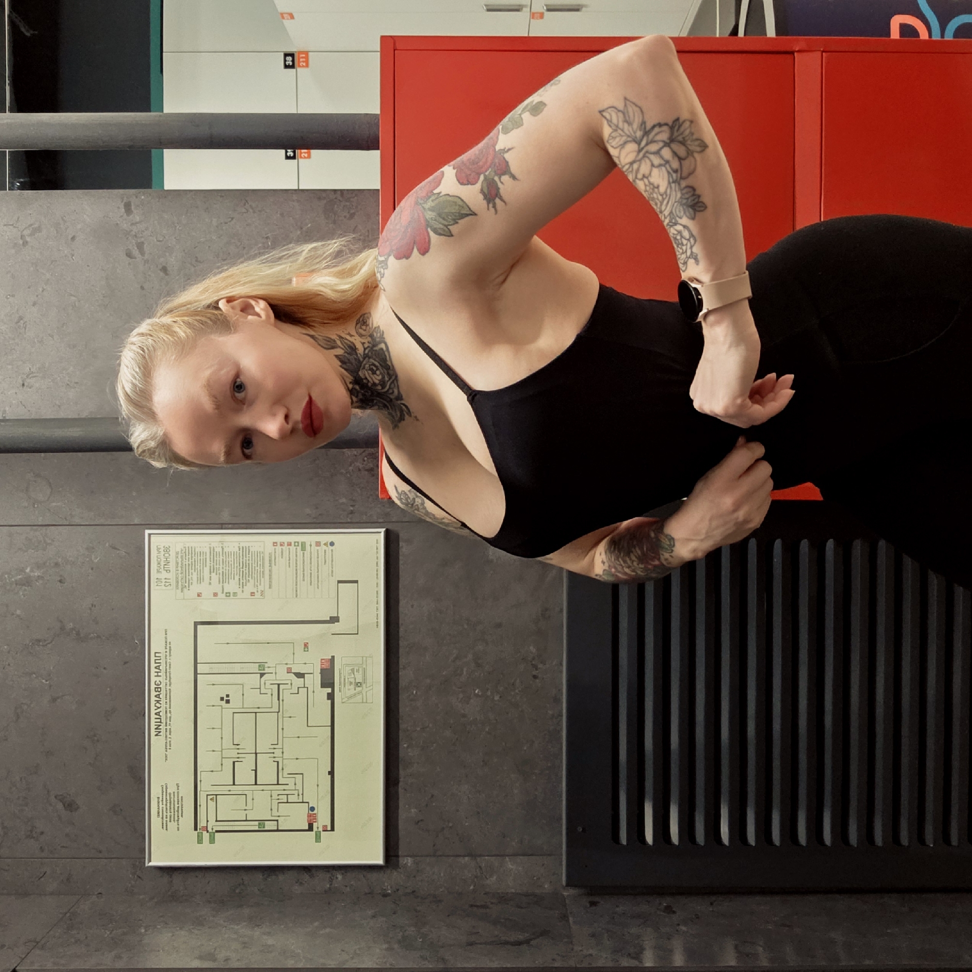 The image, rotated 90 degrees clockwise, captures a woman posing indoors, likely in a modern facility such as a gym or locker room in Saint Petersburg, Russia.
The central subject is a fair-skinned woman with blonde hair and blue eyes, wearing red lipstick. She is dressed in a form-fitting black top or bodysuit. Her left arm is raised, with her hand appearing to adjust her clothing at her side. She has a light-colored smartwatch on her left wrist. The woman is extensively tattooed; prominent floral designs are visible on her left upper arm and shoulder/neck area, and a fine-line floral tattoo adorns her right forearm. She looks directly at the camera with a neutral to confident expression.
The background features a dark grey, textured wall, possibly concrete. To the upper right, several bright red lockers are visible, some with white numbers such as "38" and "211". Below these lockers, a dark vertical grate or vent structure is integrated into the wall. On the lower left of the image, a framed diagram or map is affixed to the wall. Visible text on this document includes "NINJA VAABE HANTI" (possibly a misreading due to blur) and what appears to be Cyrillic text: "ЗВОННИК 413" (Zvonnik 413) and "UBA LUOWSE 401". The lighting is bright and even, suggesting an indoor setting with artificial illumination.