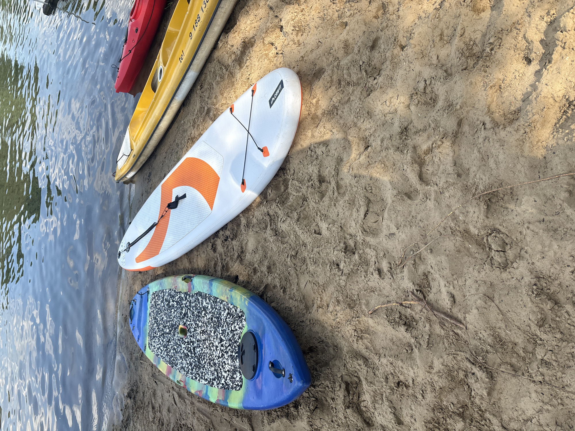 In Kobyla Góra, Poland, on a sandy shore next to a body of water, there are several watercraft lined up. A blue paddleboard with a colorful, speckled design rests on the sand, its nose pointing towards the water. To its right, a white paddleboard with an orange graphic pattern is also positioned on the sand, parallel to the blue one. Further to the right, a yellow kayak is visible, followed by a red kayak. The water exhibits ripples and reflections of the sky. The sand is coarse and shows signs of being disturbed, with small sticks and debris scattered across its surface. The scene depicts a tranquil setting by the water, with recreational equipment ready for use.