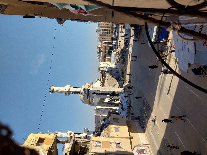 This elevated view captures a scene of extensive urban destruction in the city of Khan Yunis, Palestinian Territory, under a clear blue sky.
Central to the background is the significantly damaged Great Mosque of Khan Yunis, distinguished by its light-colored dome and a tall, slender minaret with an ornate top. Rubble and debris surround its base, indicating structural impact. To the left and right of the mosque, numerous multi-story buildings exhibit severe damage, with exposed interiors, broken facades, and piles of concrete and rebar.
In the midground, a paved street with white markings traverses the scene. Approximately 20-30 individuals, predominantly dressed in dark clothing, are walking or standing on the street and amidst the rubble. Shadows are long, extending to the left, indicating direct sunlight. On the left side of the street, a partially collapsed multi-story building features a mural depicting a figure with a raised hand on its lower section. In the lower right, a white tent with partially visible Arabic text and a blue tent are situated near the street. A person on a bicycle moves past the tents, and a figure rides a donkey-drawn cart nearby.
The immediate foreground on the right is partially obscured by the dark metal frame of a window or balcony, with light-colored laundry (pink and blue fabric) hanging. A black cable or bar extends across the lower-middle portion of the image. A faint dark line, likely an overhead utility wire, is visible against the clear sky in the upper half of the frame.