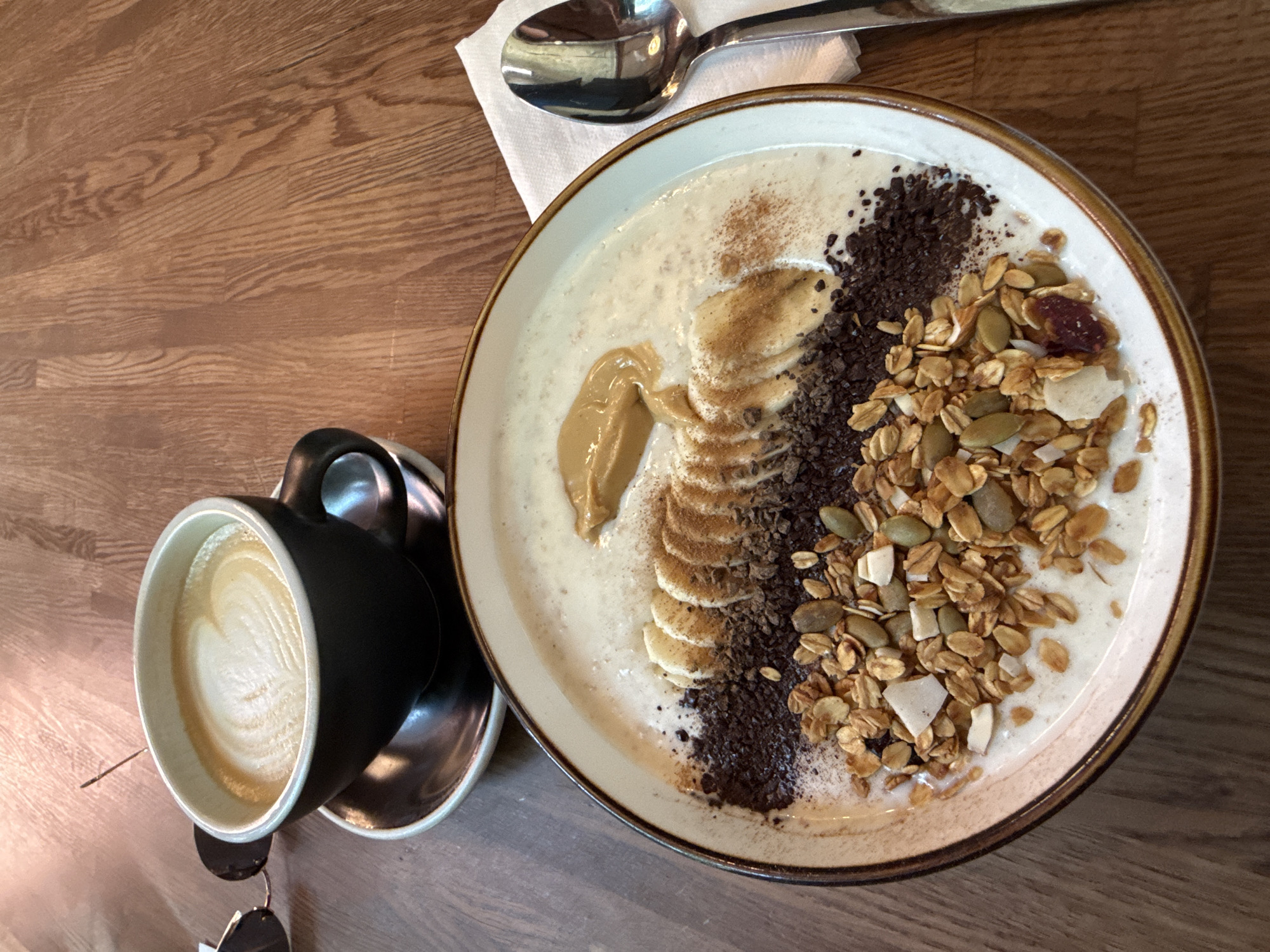 This is a close-up, high-angle shot of a breakfast meal and coffee on a wooden table. In the foreground, a bowl contains a creamy, light-colored base topped with a row of sliced bananas, a dollop of peanut butter, a scattering of dark chocolate shavings, and granola with various seeds and coconut flakes. A light dusting of cinnamon is visible on the bananas and the creamy base. To the left of the bowl, partially obscured by the bowl's rim, is a dark-colored coffee cup with a latte art design on top of the foam. The cup is on a saucer. To the left of the coffee cup are a pair of sunglasses. To the right of the bowl, a silver spoon rests on a white napkin. The background consists of a wooden tabletop with a visible wood grain pattern. The image does not provide any information about its location in Moscow, Russia.