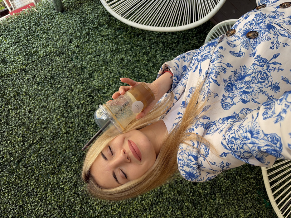 The scene is located in Budva, Montenegro. A blonde woman, approximately in her 20s or 30s, is positioned centrally in a close-up, facing slightly left with her eyes closed and a gentle smile. She is wearing a white top under a white blazer with a prominent blue floral and botanical pattern, featuring large brown buttons on the visible right side. In her right hand, she holds a clear plastic cup with a dome lid and a dark straw, containing a partially consumed iced beverage, possibly coffee, with visible residue on the interior walls. A small, white circular label is affixed to the cup. The primary background consists of a dense wall of artificial green foliage. Partially visible on the right is a section of a white, open-weave chair with a radial design, with another similar chair partially present in the lower-left foreground. The woman appears to be taking a selfie.