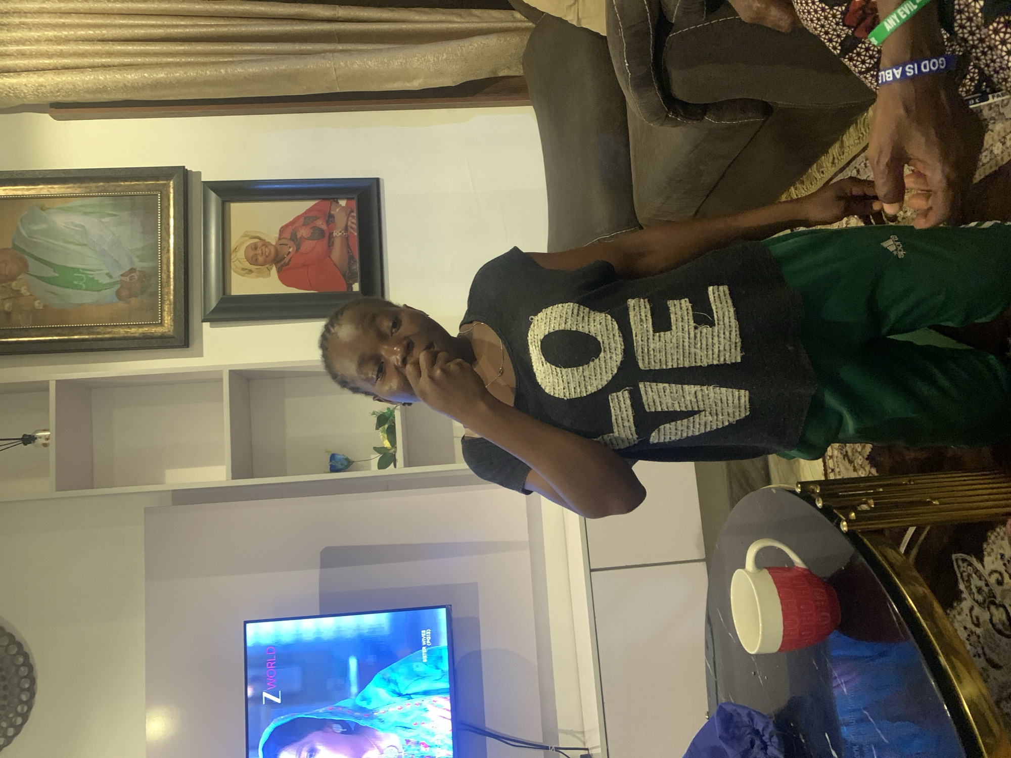 The image shows a room in a building, possibly a residential space in Jos, Nigeria. A person wearing a black top with the word "LOVE" on it and green pants is standing near a coffee table. A red and white mug is on the table. Behind the person is a white shelving unit with decorative items. On the wall above the shelving unit are two framed pictures. A television screen is visible at the bottom of the image, displaying a broadcast with an image of a person. Above the shelving unit, there is a partially visible beige curtain. To the right, another person's hand, wearing a blue bracelet with the words "GOD IS ABU," can be seen, indicating their presence in the scene. An upholstered gray chair is also visible.