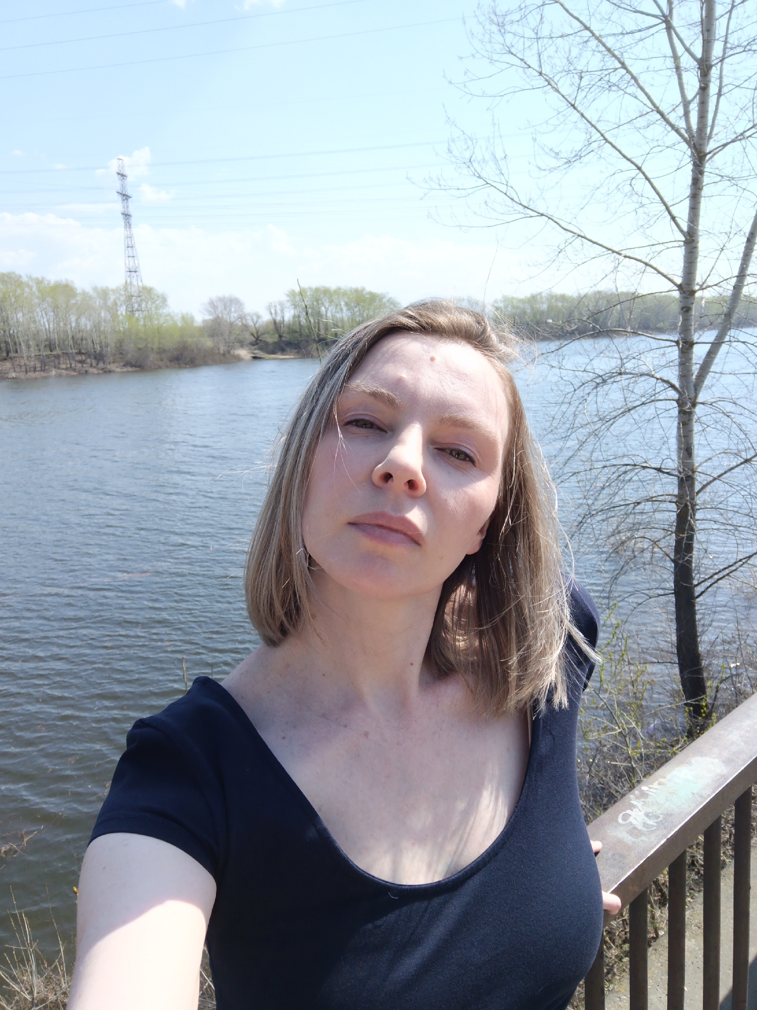 A woman is taking a selfie outdoors. The background shows a wide river with trees lining the opposite bank. In the distance, a tall electrical transmission tower is visible. A leafless tree stands to the woman's right. She is leaning against a metal railing. The sky is partly cloudy. The location is likely in the vicinity of Chaikovsky, Russia given the context.