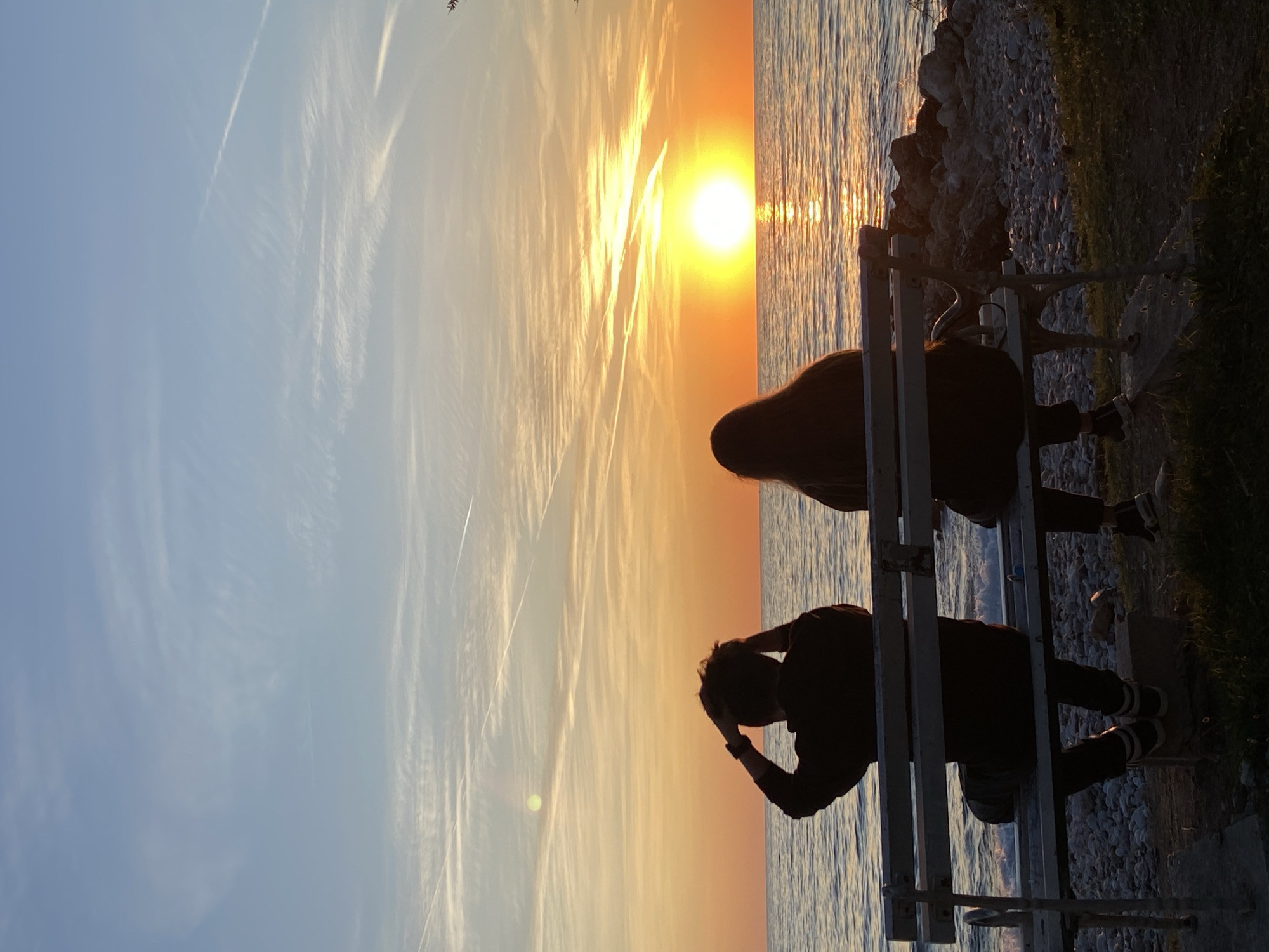 A panoramic photograph captures a serene sunset scene over a body of water in Bar, Montenegro. The image is dominated by the warm hues of late afternoon, with the sun, a bright golden orb, positioned prominently on the right side of the frame, casting a shimmering reflection across the water's surface.
Two individuals are silhouetted against the bright sky and water. They are seated on a light-colored bench, which also appears as a dark silhouette, positioned on a rocky shore. The person on the left is seen from behind, with their arm raised towards their head, as if adjusting their hair or shielding their eyes from the sun. The person on the right is also seen from behind, facing the sunset. Both figures appear to be observing the scenic view.
The sky above displays a gradient of colors, transitioning from a pale blue on the far left to vibrant oranges and yellows near the setting sun. Wispy, elongated clouds, possibly cirrus or contrails, streak across the sky, catching the golden light and adding texture to the expanse.
The foreground consists of a rocky shoreline on the right, with patches of green grass visible further in the lower right corner. The water itself appears calm, reflecting the bright sky and sun in vertical streaks of light. The overall mood of the image is peaceful and contemplative, capturing a moment of natural beauty at dusk. There is no visible text in the image.