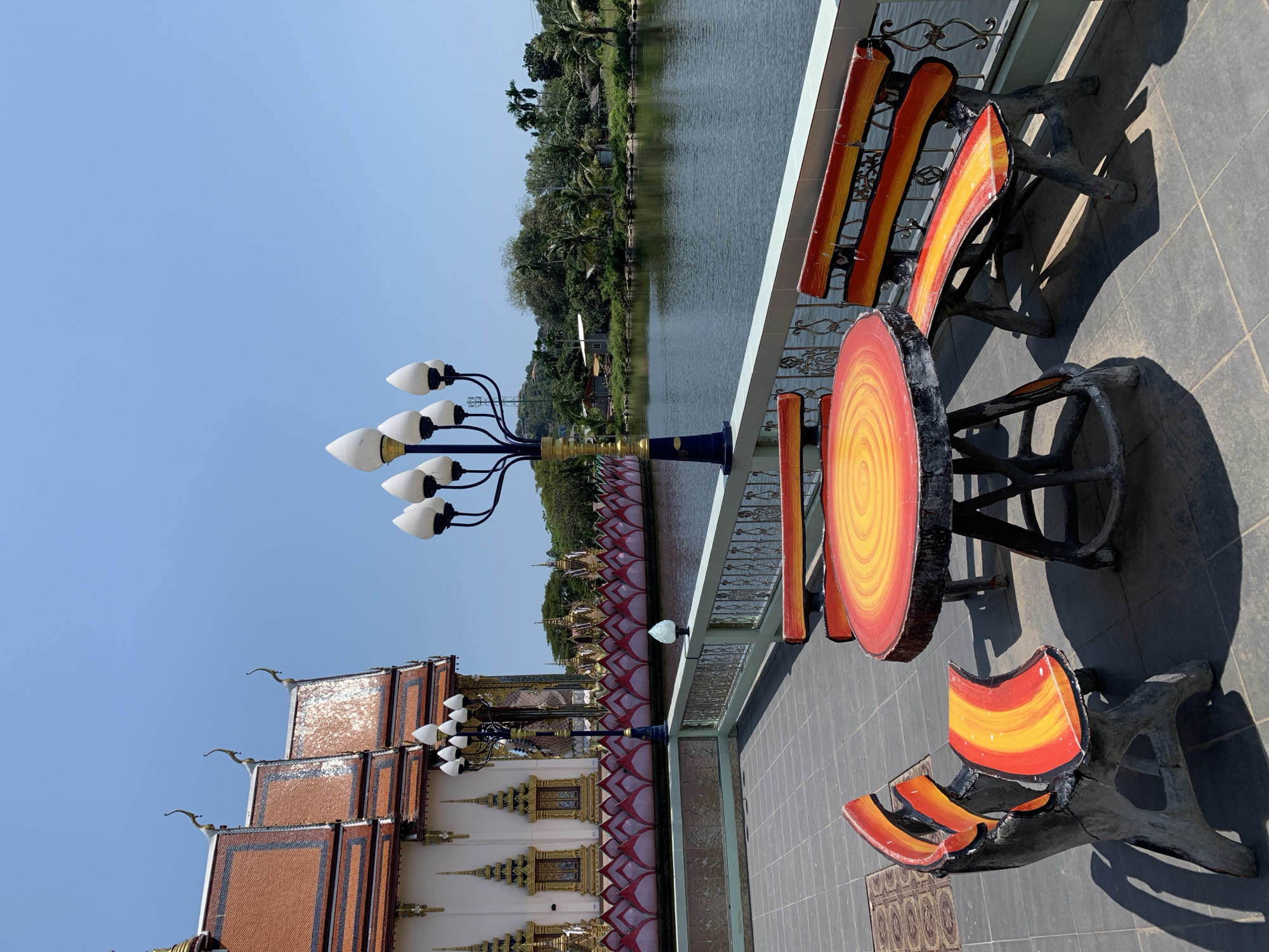 The image presents a sunlit outdoor scene situated in Baan Plai Laem, Thailand. In the immediate foreground, a grey tiled patio floor extends. Centrally positioned on the patio is a concrete table with a circular top, featuring concentric rings of red, orange, and yellow paint on a dark grey base. Two matching concrete benches, painted with red, orange, and yellow stripes on their seats and backs, flank the table; one is partially visible on the far left, and another on the far right. Strong shadows from the furniture are cast onto the patio. Behind the furniture, a decorative street lamp with a blue and gold pole supports seven white, lotus-bud shaped light fixtures arranged on black arms.
A light blue and grey railing with black wrought iron scrollwork separates the patio from a body of calm, dark water, indicative of a lake or lagoon. To the left, a traditional Thai temple building with white walls, a multi-tiered terracotta-tiled roof, golden decorative elements, and ornate window frames stands prominently. Along the water's edge, adjacent to the temple, a continuous border of floating or built structures resembling pink and red lotus flowers stretches across the surface. The background on the right side of the water reveals a dense, tropical landscape of lush green foliage, numerous palm trees, distant hills, and a few small, indistinct buildings. The sky above is clear and uniformly blue. No individuals or active movements are observed.