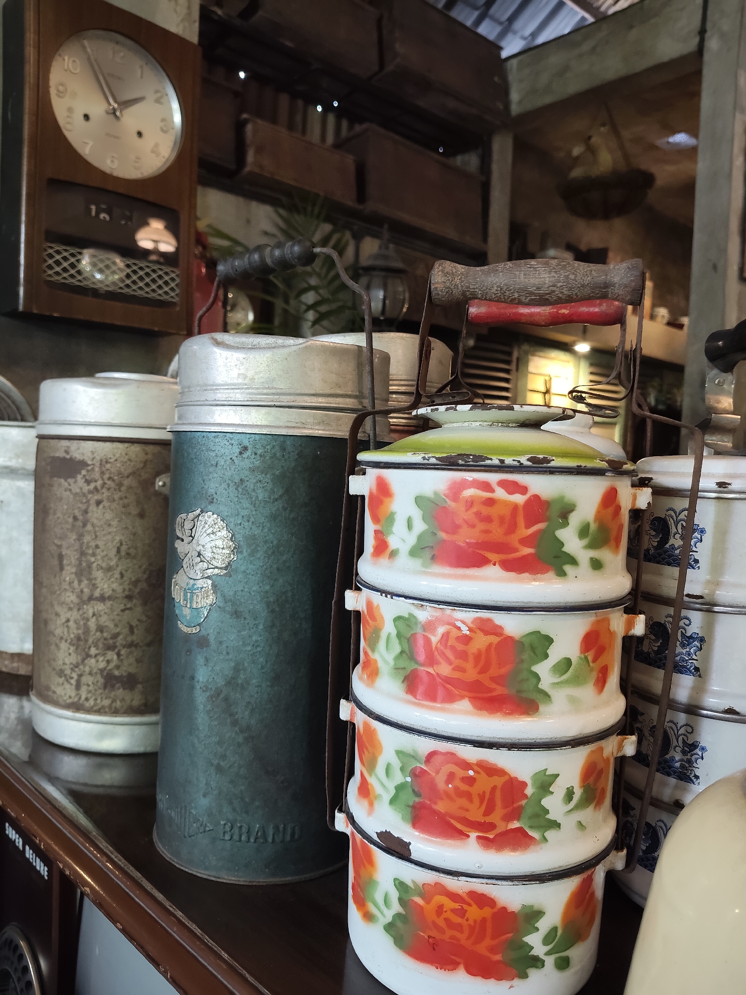 The image depicts a collection of vintage kitchenware items displayed on a dark brown shelf in what appears to be a cafe or antique shop in the city of Medan, Indonesia. The primary focus is on a stack of three enamel tiffin carriers, each decorated with hand-painted red and orange roses and green leaves against a white background. The tiffin carriers have metal handles and are supported by a rusty metal frame. To the left of the tiffin carriers are two vintage thermoses or canisters. The taller, teal-colored thermos has a faded emblem with an image of a shell and text that appears to read "COLUMBIA BRAND". The shorter, left thermos is a mottled brown and silver color, showing signs of age and rust. Behind the thermoses, a wooden-cased clock with a silver face hangs on the wall, displaying the time as approximately 10:10. The background suggests an indoor environment with a corrugated metal ceiling and exposed beams, characteristic of older buildings in Southeast Asia. The overall scene evokes a sense of nostalgia and is likely a curated display of antique items.