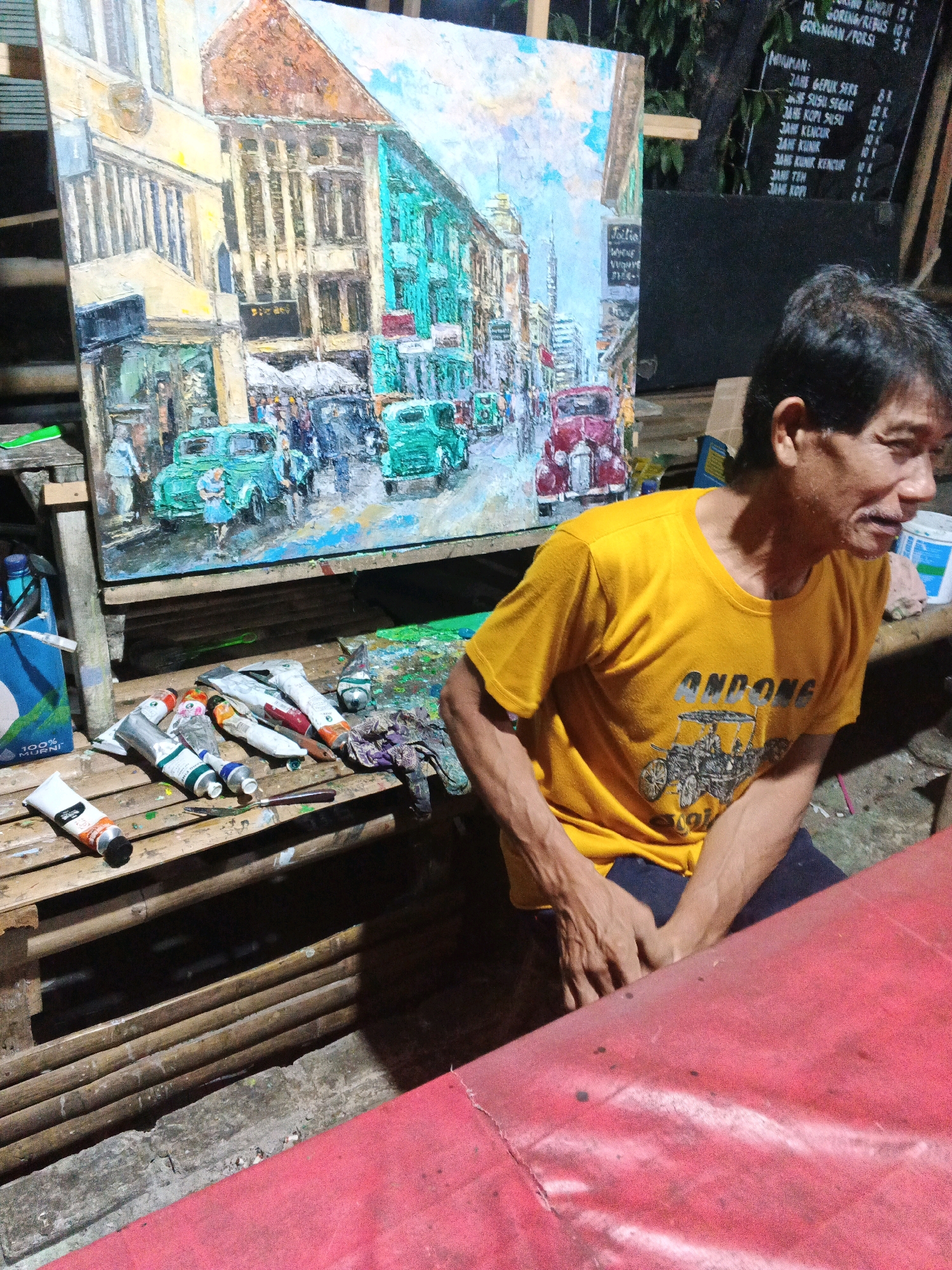 An adult male with dark hair and a yellow t-shirt is seated in front of a painting on an easel. The painting depicts a street scene with buildings and vintage cars. Tubes of paint and art supplies are laid out on a wooden surface. A red surface is visible in the foreground. The background includes greenery and a blackboard with text. The man appears to be an artist in Ungaran, Indonesia, possibly at a market or outdoor studio.