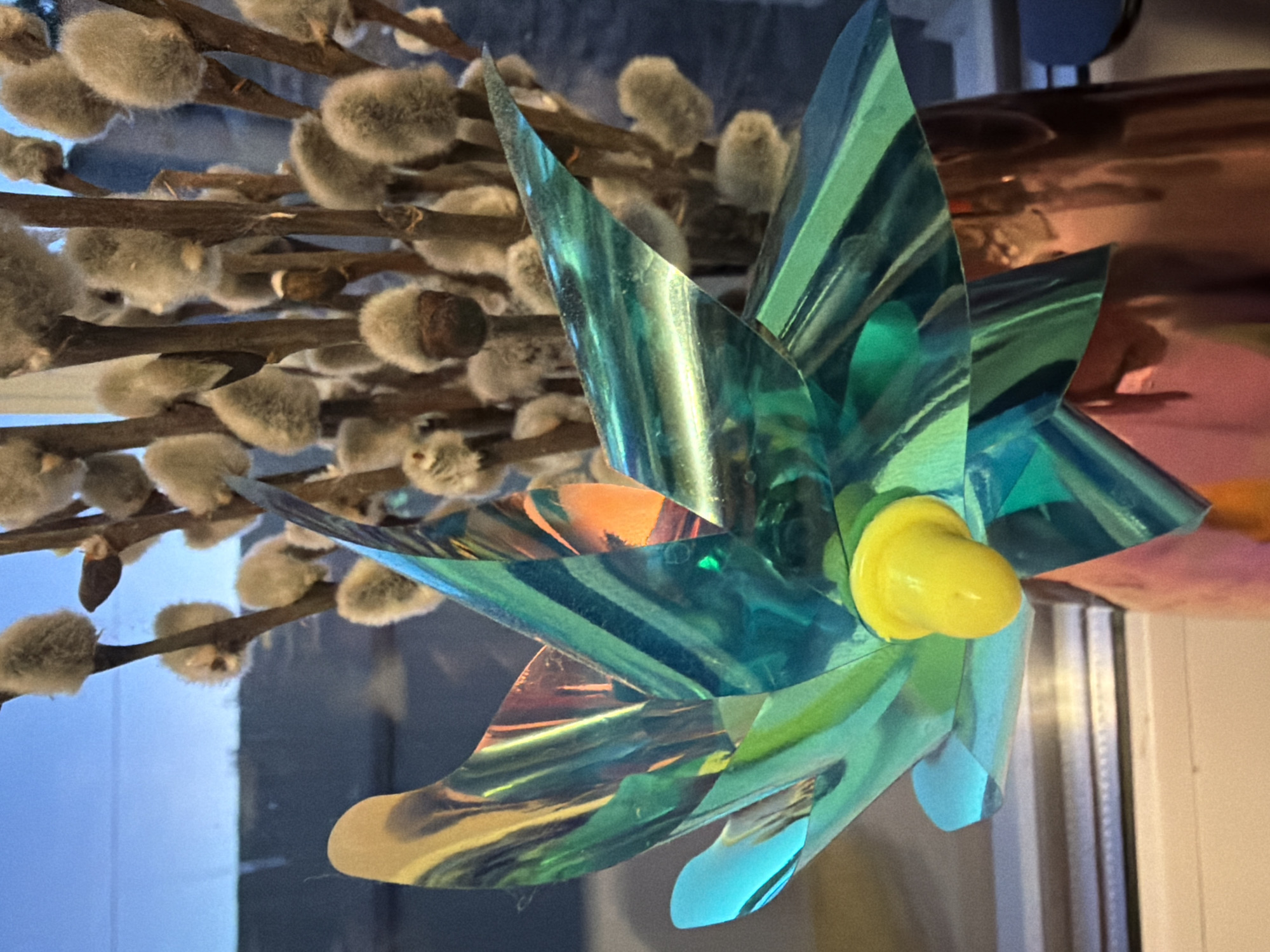 A still life photo shows a close-up view of a decorative pinwheel with iridescent green and blue blades, topped with a yellow pom-pom, resting in a copper vase filled with pussy willow branches. The background is a window showing a blurred view of an outdoor scene with a blue sky. The lighting is soft, suggesting it might be early morning or late evening. The overall composition is balanced, with the pinwheel as the main subject. The scene does not depict any specific location in Bila Tserkva, Ukraine, as there are no identifiable landmarks or context clues present in the image.