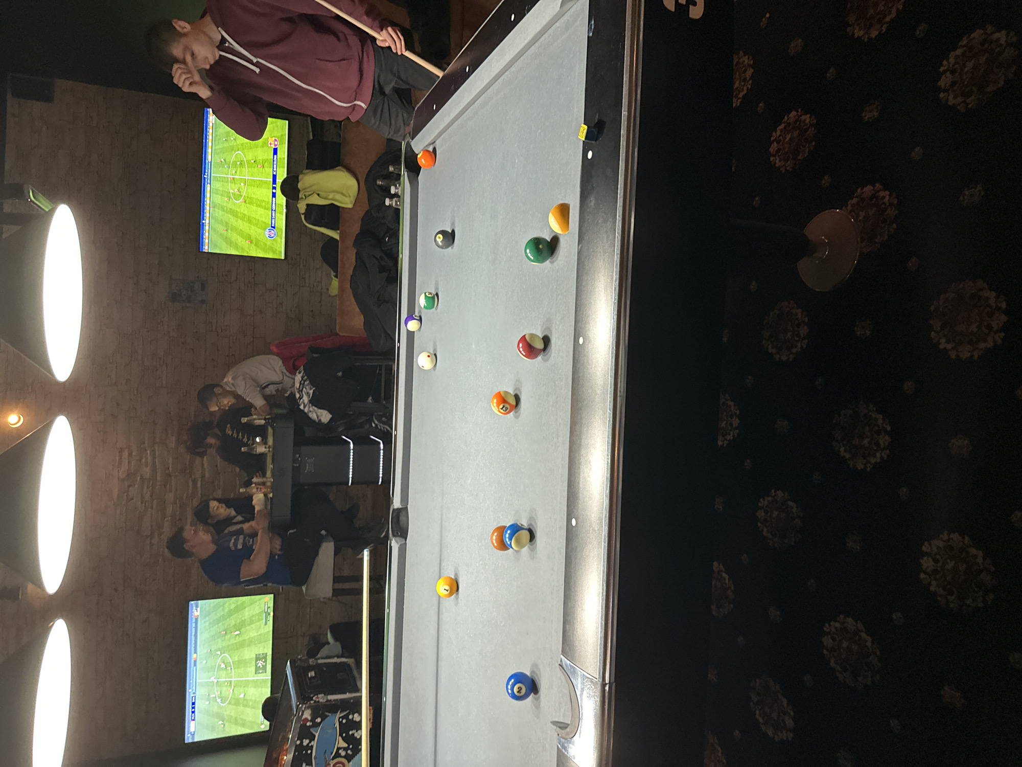 The image shows the interior of a bar or pub. A billiard table dominates the foreground, with multiple billiard balls scattered across its surface. A person is standing at the head of the table, holding a pool cue and apparently preparing to take a shot. In the background, several people are seated at a table. Two television screens are visible, displaying a football match. The walls are decorated with brickwork and patterned wallpaper. The lighting is provided by multiple spotlights mounted on the ceiling. There is no explicit indication that the location is in Plovdiv, Bulgaria.