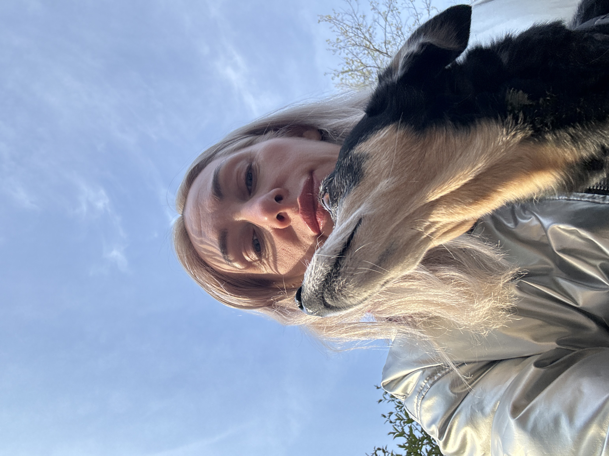 A low-angle, close-up shot shows a blonde woman in a silver jacket taking a selfie with a dog. The woman is on the left side of the frame and is looking down at the camera. The dog is on the right side of the frame and is nudging its snout against the woman's face. The background is a blue sky with some light clouds. The shot could have been taken anywhere, with no indication of it being in Saint Petersburg, Russia.