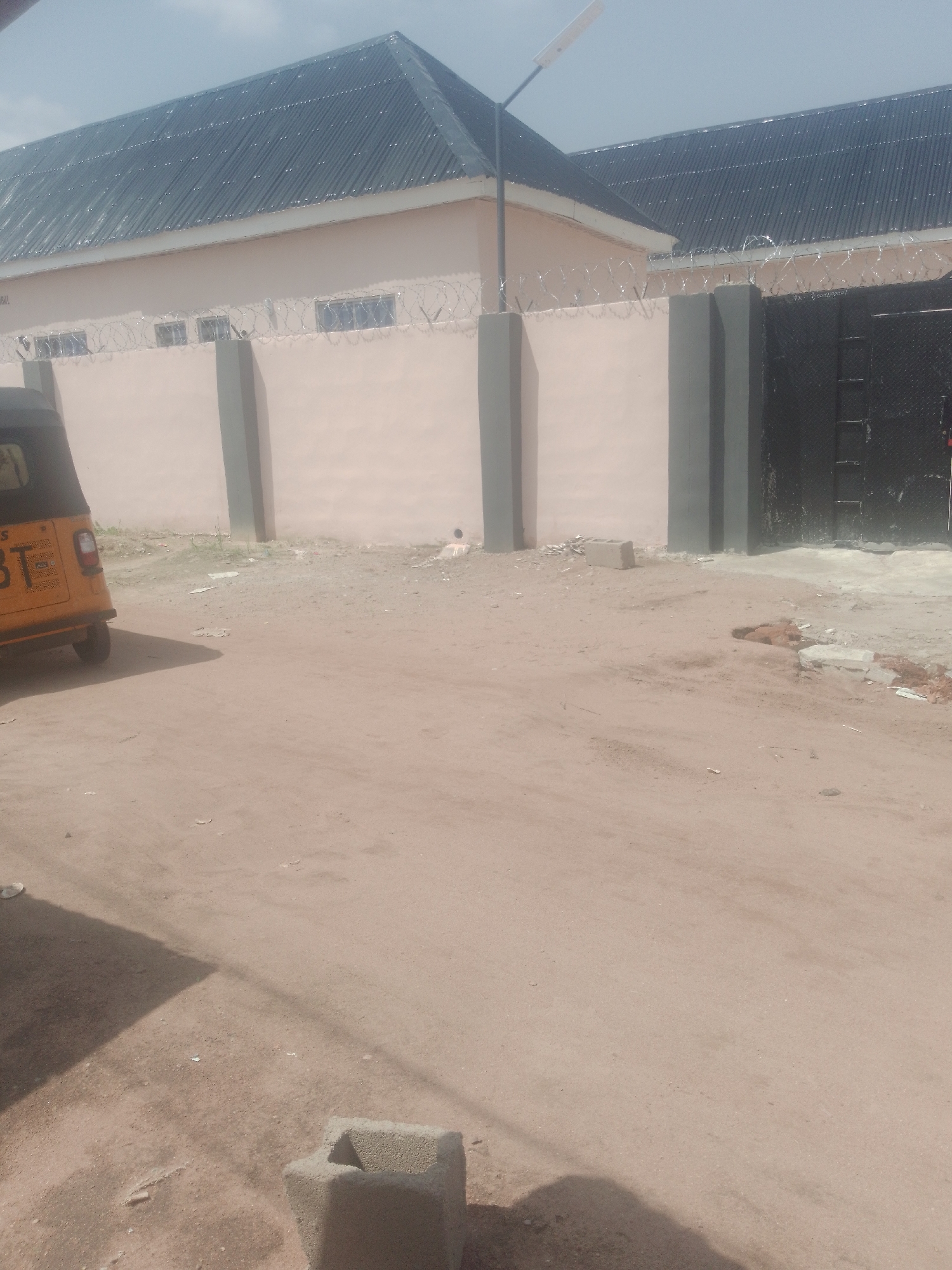The image depicts a scene in Jauro Yinu, Nigeria, featuring a light pink building with a dark corrugated metal roof. A tall black metal gate is visible on the right side of the building, secured by barbed wire along the top of the wall. In front of the building, there is a sandy ground with some scattered debris. To the left of the building, a yellow three-wheeled vehicle, commonly known as a Keke Napep, is parked. The sky is overcast with light clouds. The overall impression is that of a modest building with some security features in a semi-urban or rural setting in Nigeria.