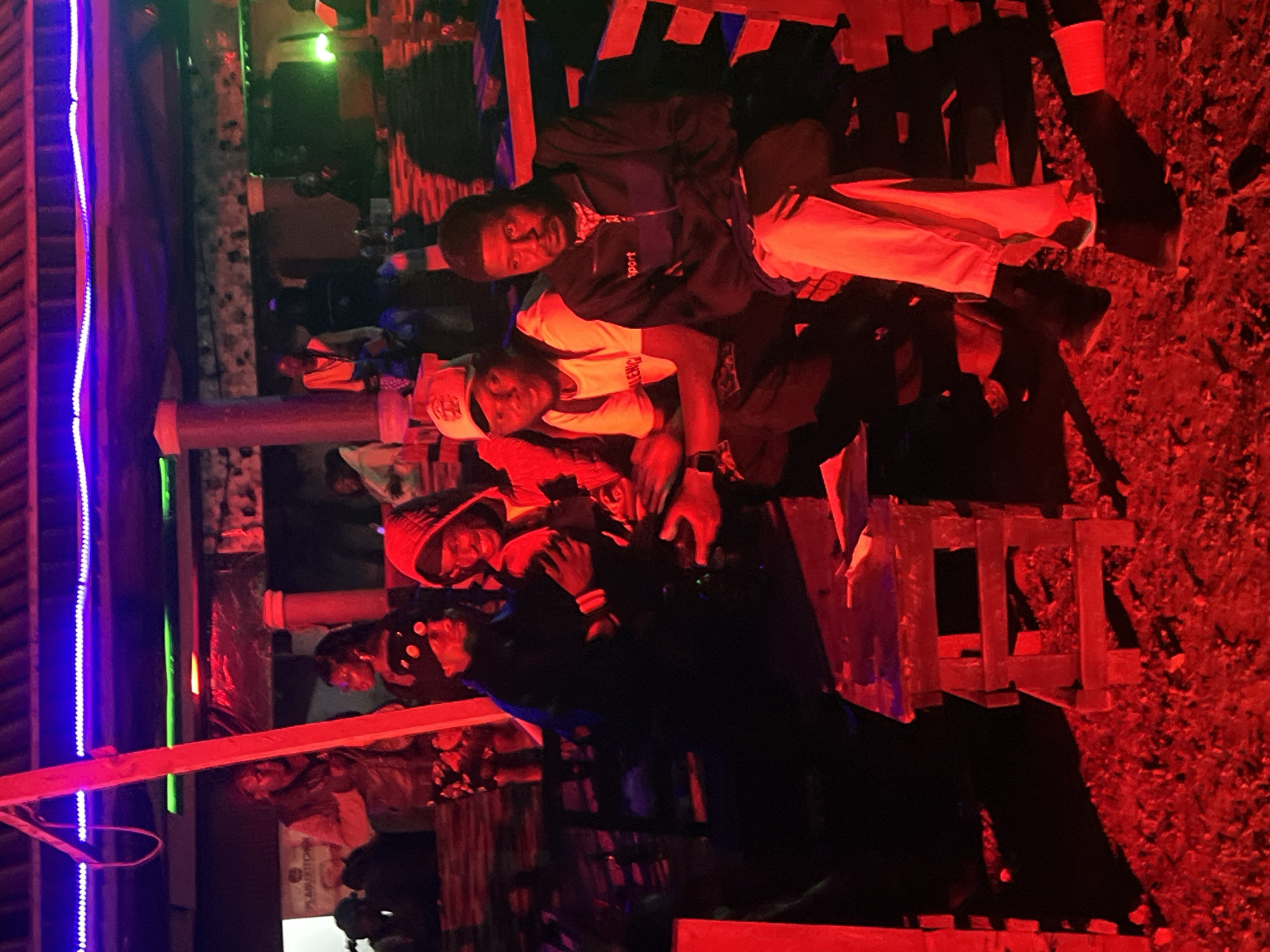 A group of young men are gathered at an outdoor venue in Durumi, Nigeria. The scene is illuminated by red and purple neon lights, creating a vibrant and somewhat diffused atmosphere. In the foreground, two men are seated on a wooden bench. The man on the left wears a white t-shirt with lettering and a light-colored baseball cap. The man on the right is wearing a dark zip-up jacket with "Sport" visible on it and has his arms crossed. Behind them, other individuals are visible, some seated and others standing. There's a wooden structure in the middle ground, possibly a table or a makeshift stand, and the ground appears to be covered in gravel or rough terrain. The overall impression is that of a casual gathering or social event at night.
