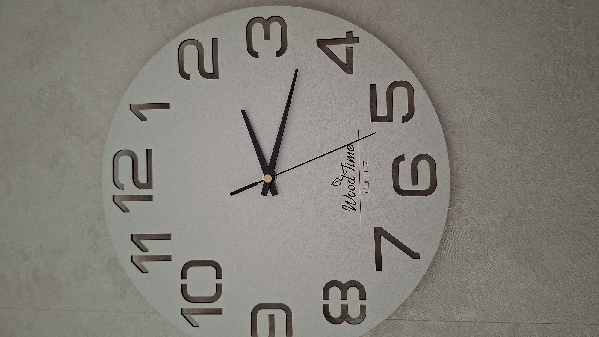 A light-colored, round wall clock is prominently displayed on a textured, off-white wall, captured from a low angle. The clock features large, dark, cut-out numerals from 1 to 12. The numbers 1, 2, 3, 4, 5, 6, 7, 8, 10, 11, and 12 are visible, while number 9 is mostly out of frame.
The clock has three black hands emanating from a central gold-colored pin. The hour hand points just past the 11, and the minute hand points directly at the 2, indicating the time is approximately 11:10. A thin second hand points towards the 5.
Text printed on the clock face reads "Wood Time" in an elegant script font, accompanied by a small leaf or branch icon, and "QUARTZ" in a smaller, sans-serif font below it. The wall behind the clock has a subtle, uneven texture, suggesting wallpaper or a specialized paint finish. The overall aesthetic is clean and modern.
