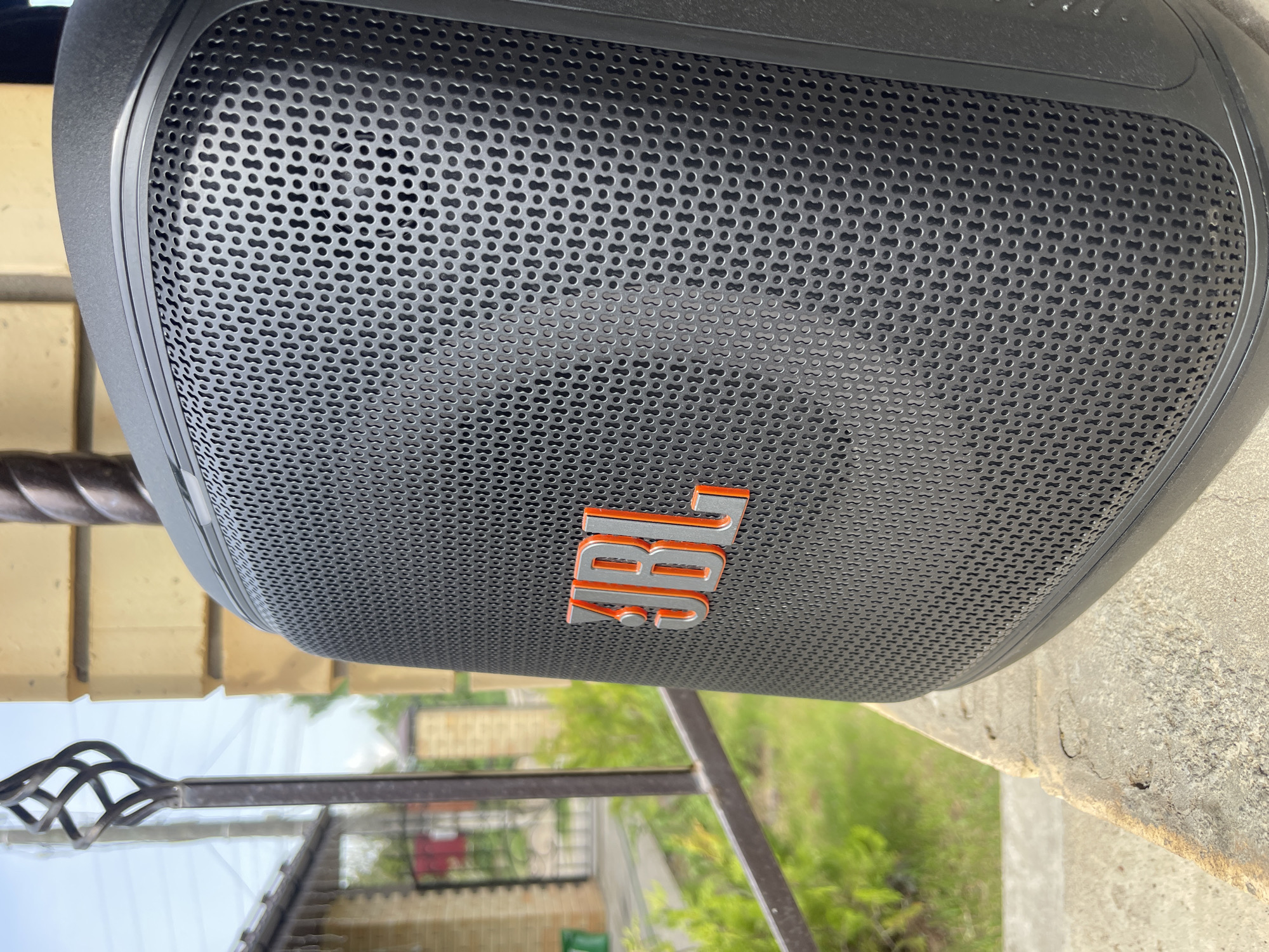 The image shows a close-up of a black JBL speaker with an orange logo. The speaker is situated outdoors, likely on a patio or balcony, based on the view of greenery and architectural elements in the background. A metal railing is visible in the lower part of the image, as well as a brick structure to the left and concrete to the right of the speaker. While the location is specified as the city of Chaikovsky, Russia, the image itself doesn't provide any visual details to confirm this information.