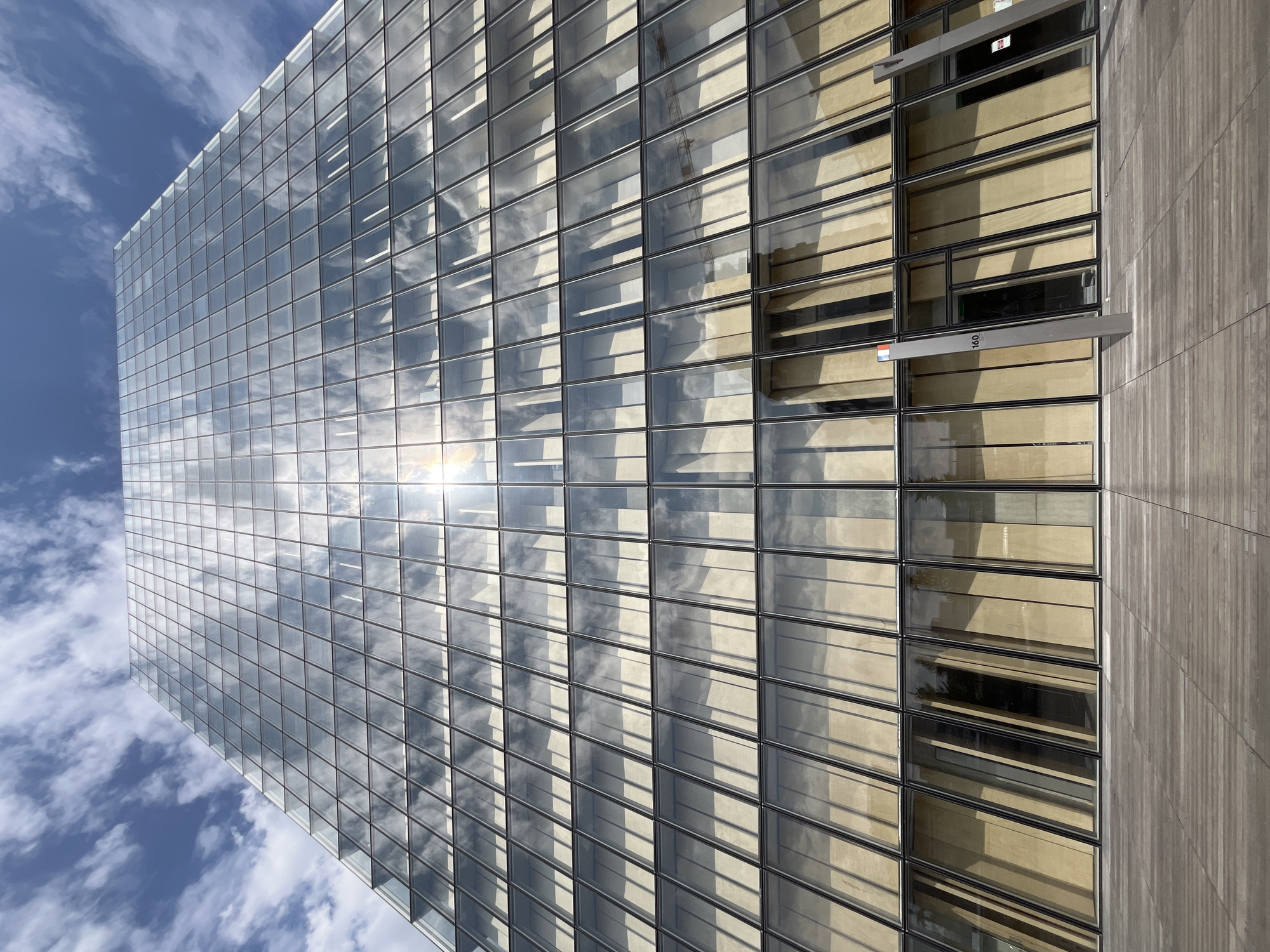The media file depicts the exterior of a modern glass-clad skyscraper on a sunny day. The building's facade is composed of numerous rectangular glass panels, reflecting the bright blue sky and scattered white clouds. The sun is prominently visible, shining directly on the glass and creating a strong glare. On the right side of the building, a section of the facade is made of a light-colored material, possibly wood or stone, with vertical lines creating a textured appearance. Some of the glass panels appear to be openable windows or vents.
Notable details include the reflection of clouds and the sun on the glass, which creates a dynamic visual effect. Shadows are cast on the building, indicating the time of day is likely morning or afternoon. There are a few small pieces of text visible on the building: a red and white sticker on one of the upper window frames, and the number "160" on a silver protrusion near the lower right section of the building, accompanied by a small Dutch flag. The overall impression is one of architectural modernity and a clear, bright day. No people or specific activities are discernible.
