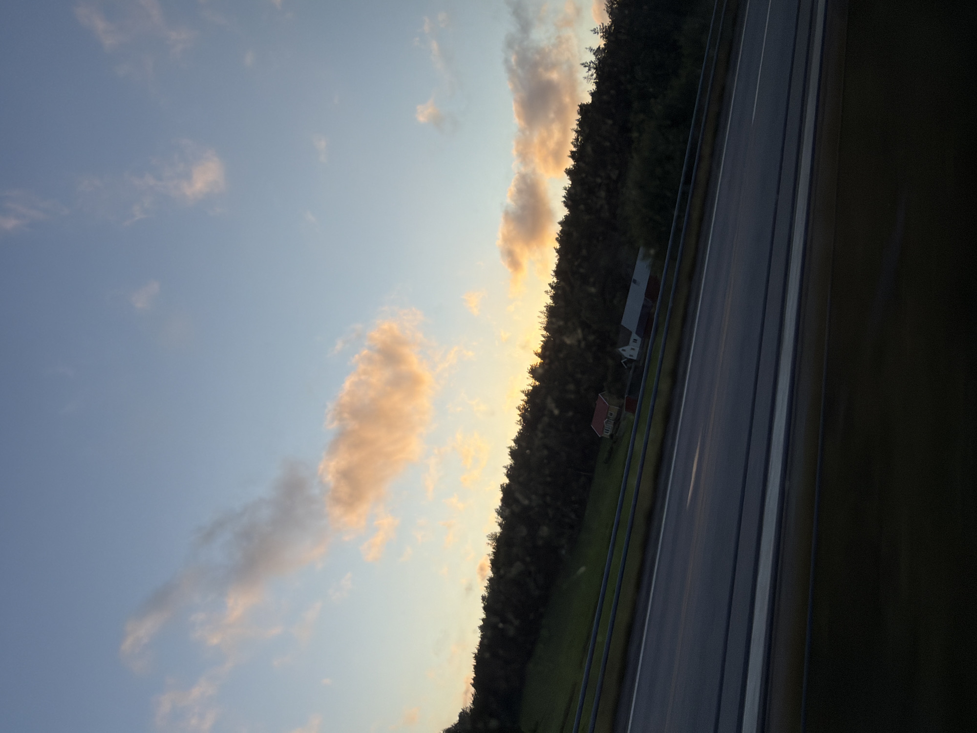 The image depicts a scenic view with a clear blue sky and scattered clouds tinged with orange and pink hues, suggesting either sunrise or sunset. A hillside covered in dense, dark green trees slopes upwards from a highway. On the hillside, two small buildings are visible: a red-roofed structure and a white building with a darker roof. The highway in the foreground has multiple lanes and is bordered by dark railings. The overall impression is of a peaceful, rural landscape. There are no people visible in the image. The location is identified as Rabbalshede, Sweden, although this cannot be independently verified from the visual information provided.