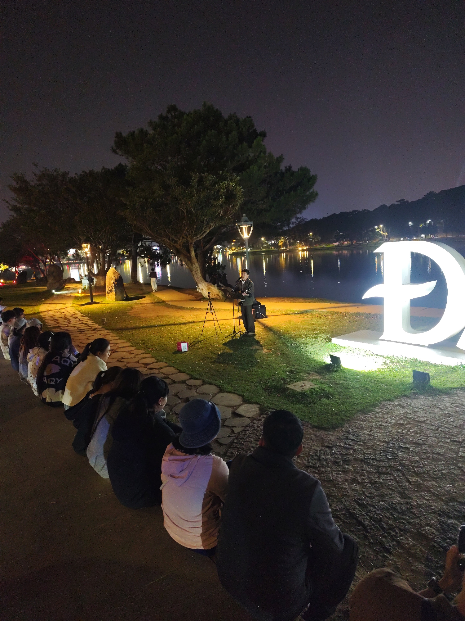 The image depicts an intimate outdoor musical performance or gathering taking place at night in Da Lat, Vietnam. A group of people, primarily young adults, are seated on the ground along a stone-paved path, attentively watching a performer. The performer, dressed in dark clothing, stands illuminated by artificial lights, playing an acoustic guitar and singing into a microphone. A speaker and tripod are positioned near the performer.
The setting is a grassy bank beside a calm lake, which reflects the numerous city lights and streetlights from the opposite shore, creating a luminous effect on the water. Large trees with sprawling branches are visible, and traditional streetlights cast warm light onto the scene. A prominent, large, white, illuminated letter "Đ" stands on the grass near the lake, serving as a clear visual cue for Da Lat. The sky is dark, indicating nighttime, and the artificial lighting focuses attention on the performer and the immediate surroundings. The atmosphere appears peaceful and engaging, with the audience focused on the performance.