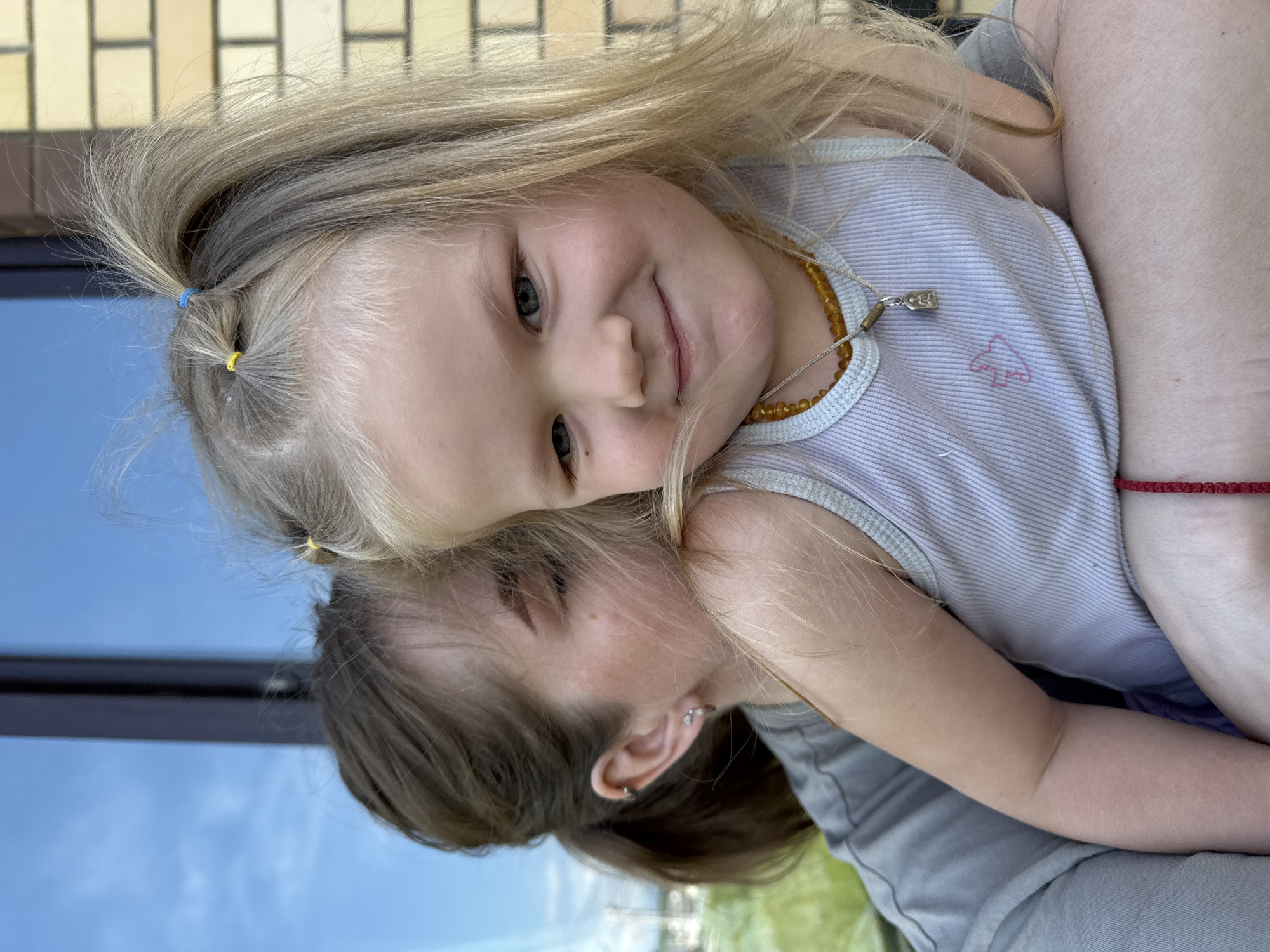 A young blonde girl with blue and yellow hair ties and an amber necklace is being held by a woman. The girl is wearing a striped tank top with a small pink design. The woman has brown hair and earrings. They are outside in what appears to be Ферма, Russia, near a building with reflective windows that show the sky.