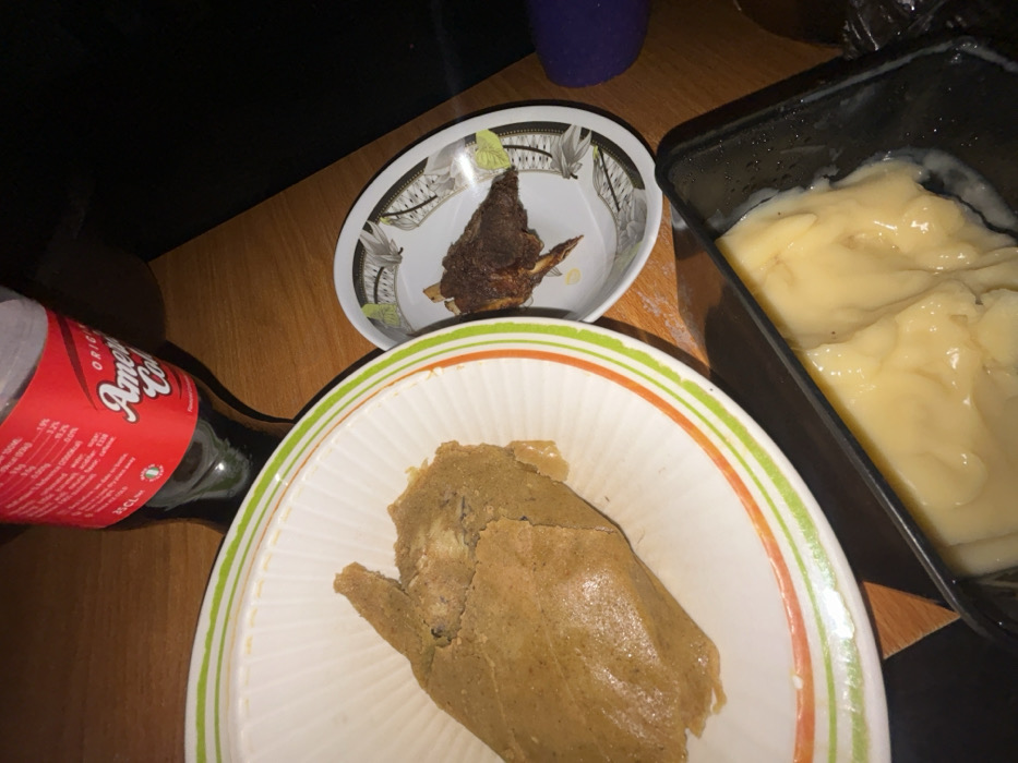 The image depicts an indoor meal setup, reportedly located in Debasco, Nigeria. No individuals are visible within the frame. The primary subjects are various food and beverage items arranged on a light brown wooden surface.
In the upper central portion, a clear plastic bottle containing a dark liquid, identified by its red label with white text as "America Cola" and "25 CL" volume, stands upright. To the left, a large, irregularly shaped mass of light brown, cooked food, consistent with a steamed pudding or paste (e.g., *moin-moin*), is presented in a white ceramic bowl with distinct green and orange stripe patterns along its rim. To the right of this bowl, a smaller, dark brown piece of cooked meat, possibly bone-in and appearing charred or fried, rests within a shallow white ceramic bowl adorned with a black and white geometric pattern and green leaf motifs. In the lower right foreground, a black rectangular plastic container holds a viscous, pale yellow food substance, resembling a starch-based swallow (e.g., *fufu* or *amala*) or a creamy pudding, with some liquid residue visible along its interior edges. The scene is illuminated, suggesting an ambient light source from the left or above.